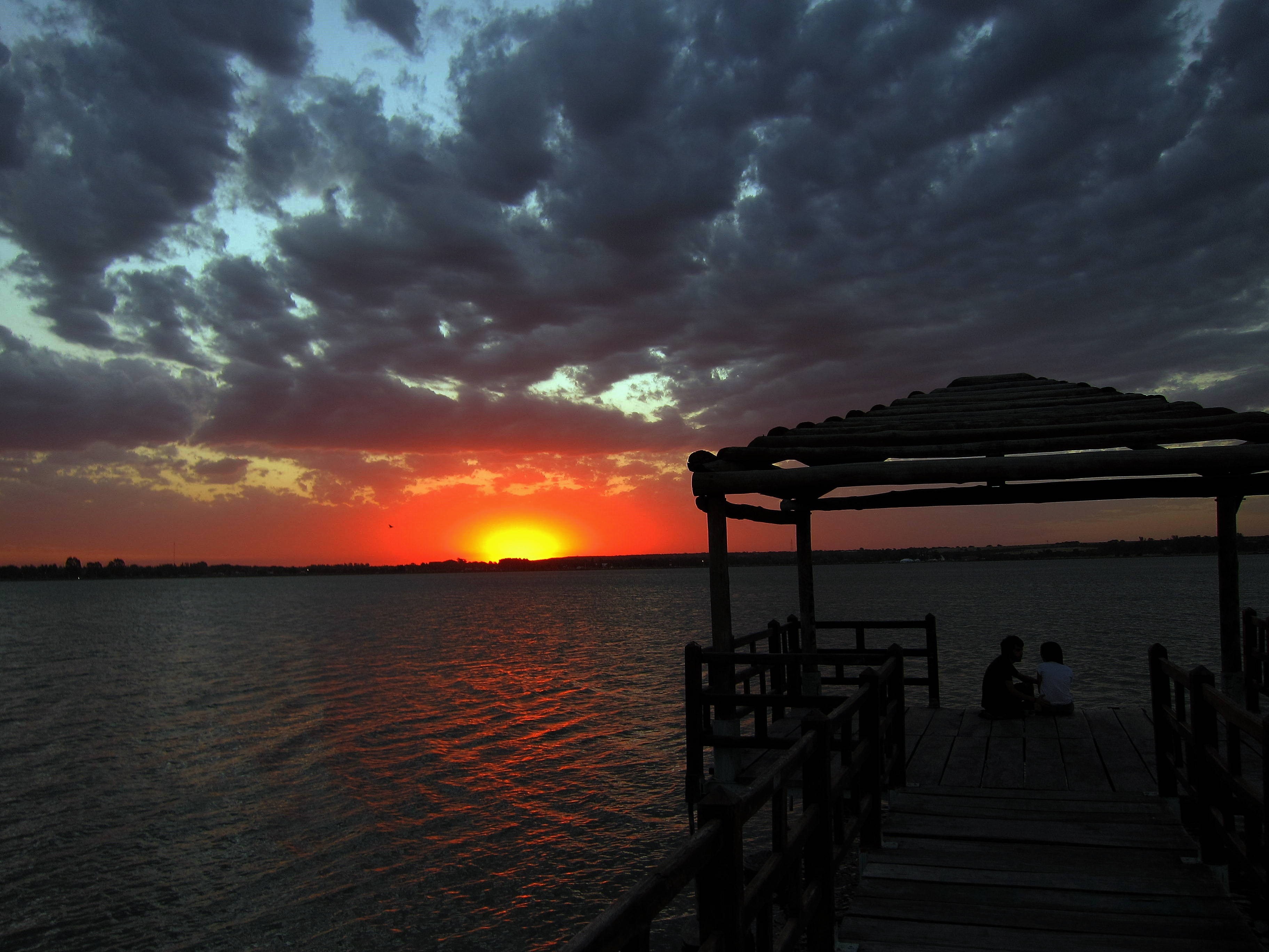 Two children on a pier watching a dramatic sunset over the ocean HD Wallpaper