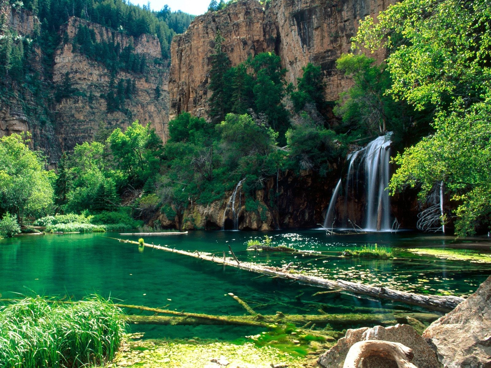 Waterfall cascading into a clear emerald lake surrounded by cliffs and trees HD Wallpaper