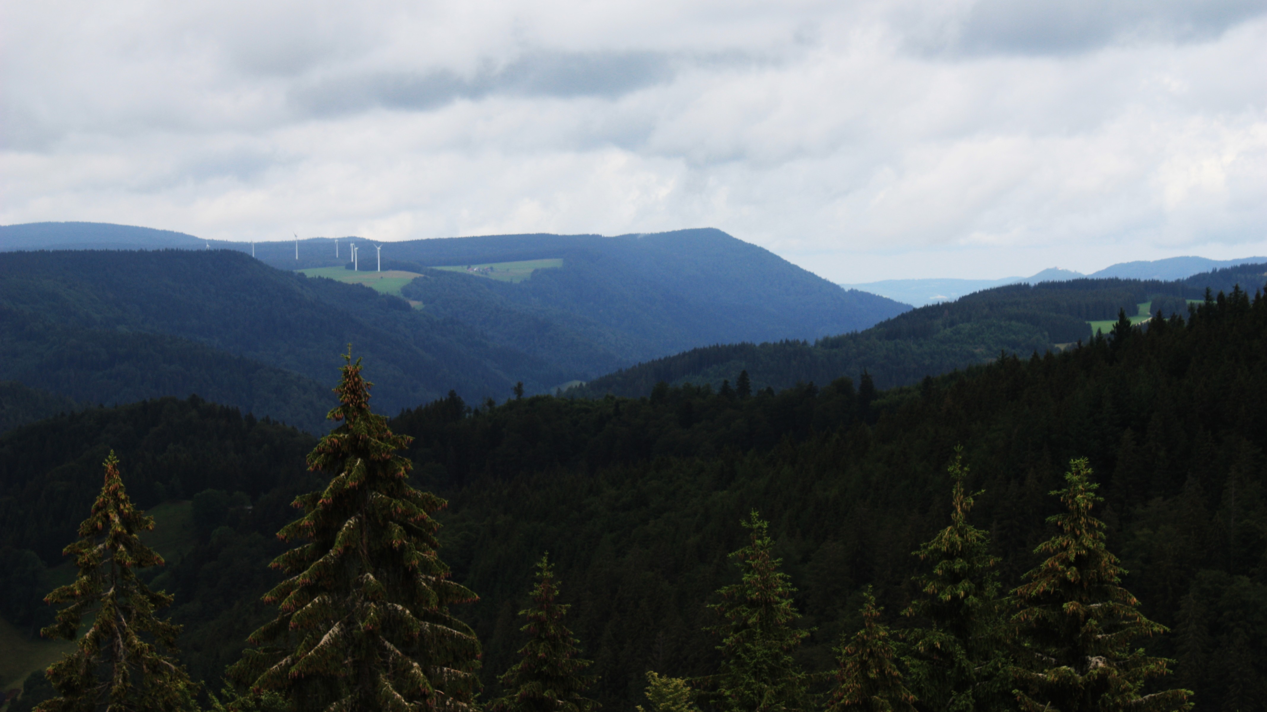 Wind turbines on a distant hill overlooking a forested mountain range HD Wallpaper