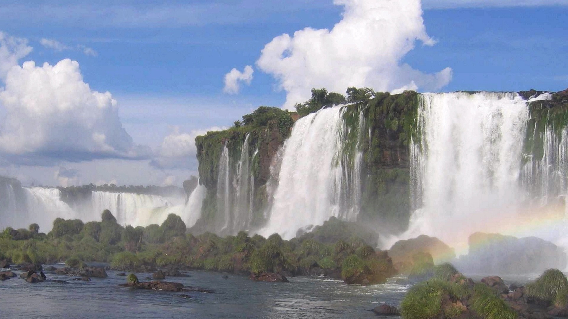 A wide waterfall cascades over a cliff with lush vegetation HD Wallpaper