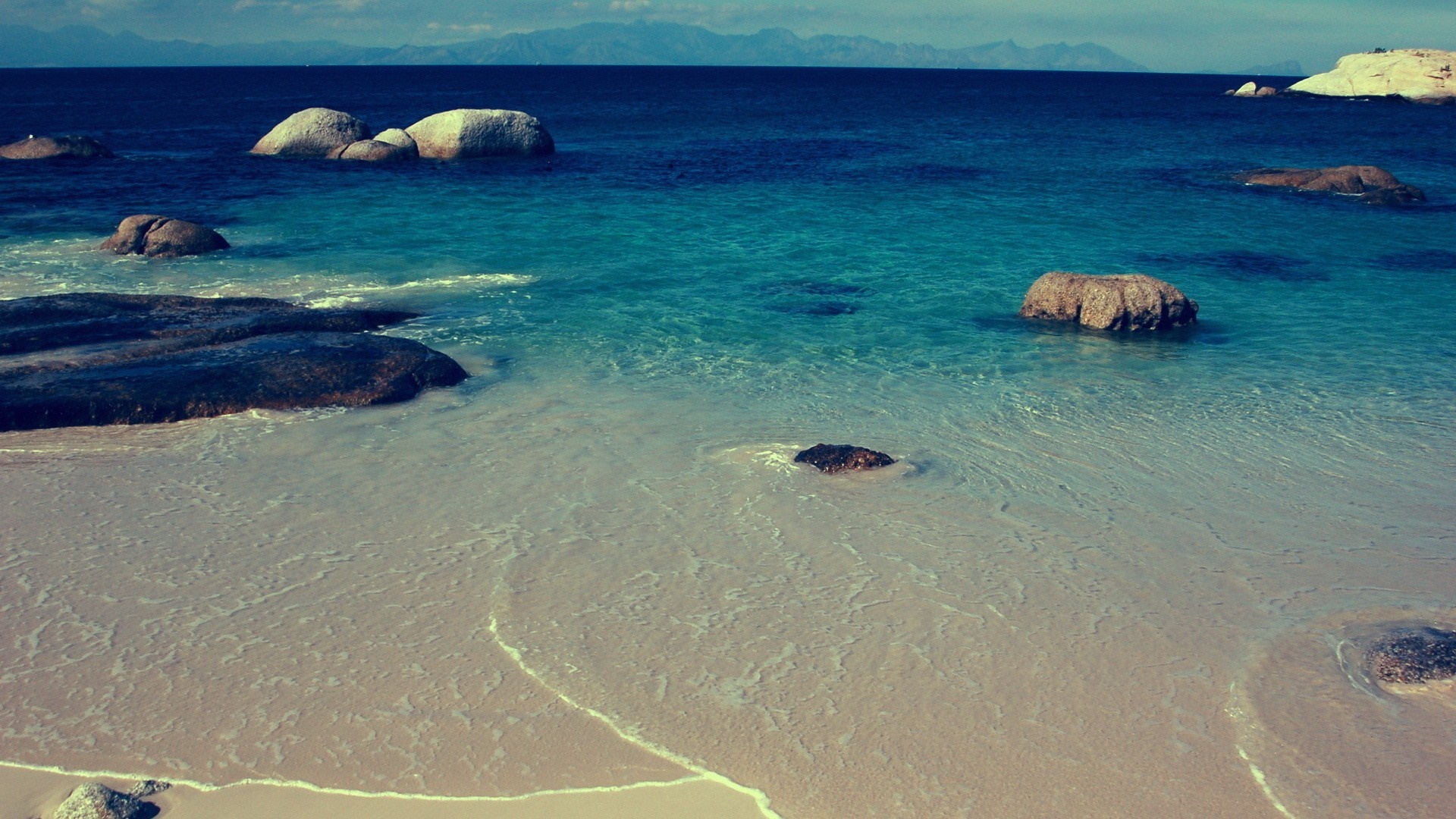 Ocean waves lap on a sandy beach with large rocks HD Wallpaper