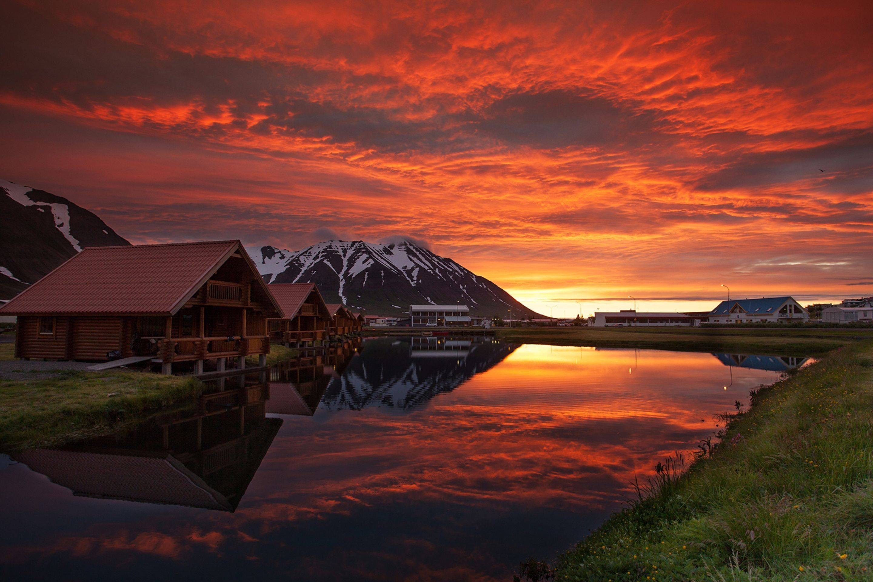Wooden cabins by a lake during a vibrant sunset with mountains HD Wallpaper