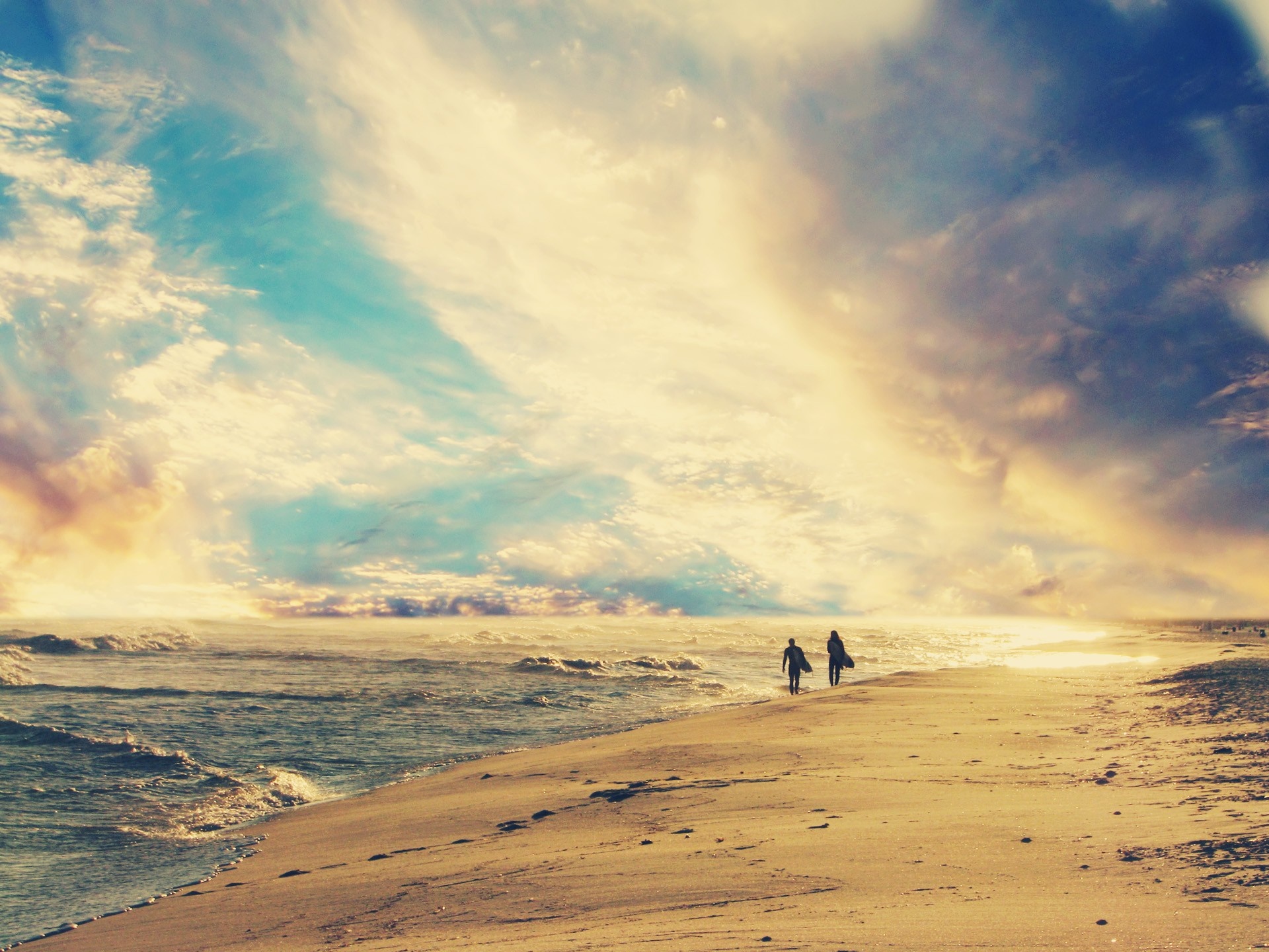 Two surfers walk along a sandy beach at sunset HD Wallpaper