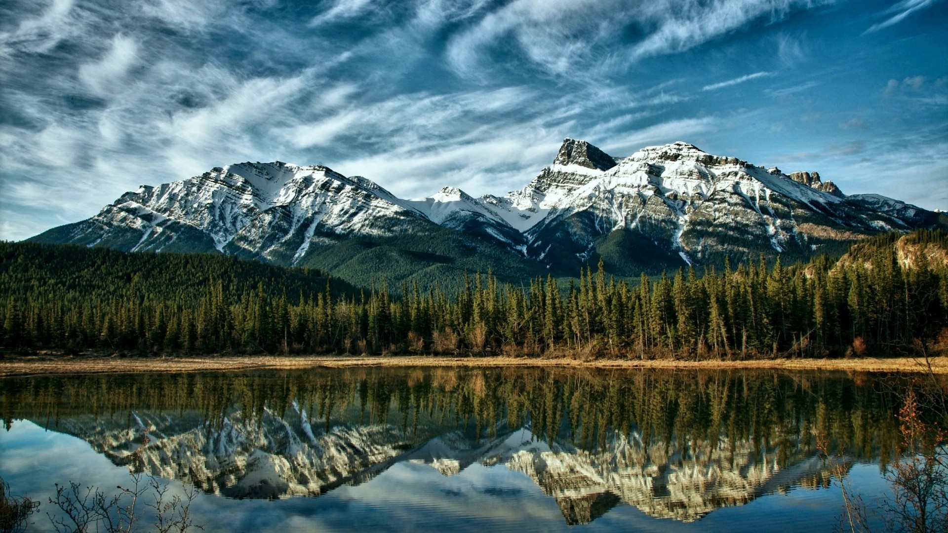 Snow-capped mountains reflected in a calm lake with pine trees HD Wallpaper