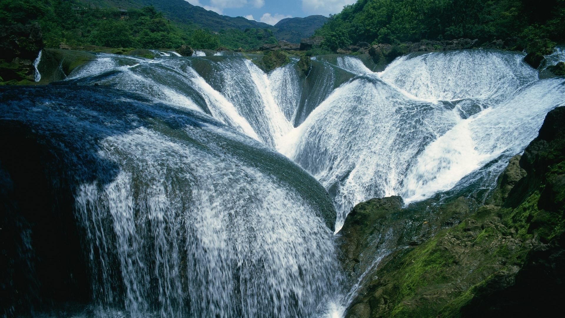 A wide, multi-tiered waterfall cascades over moss-covered rocks. HD Wallpaper