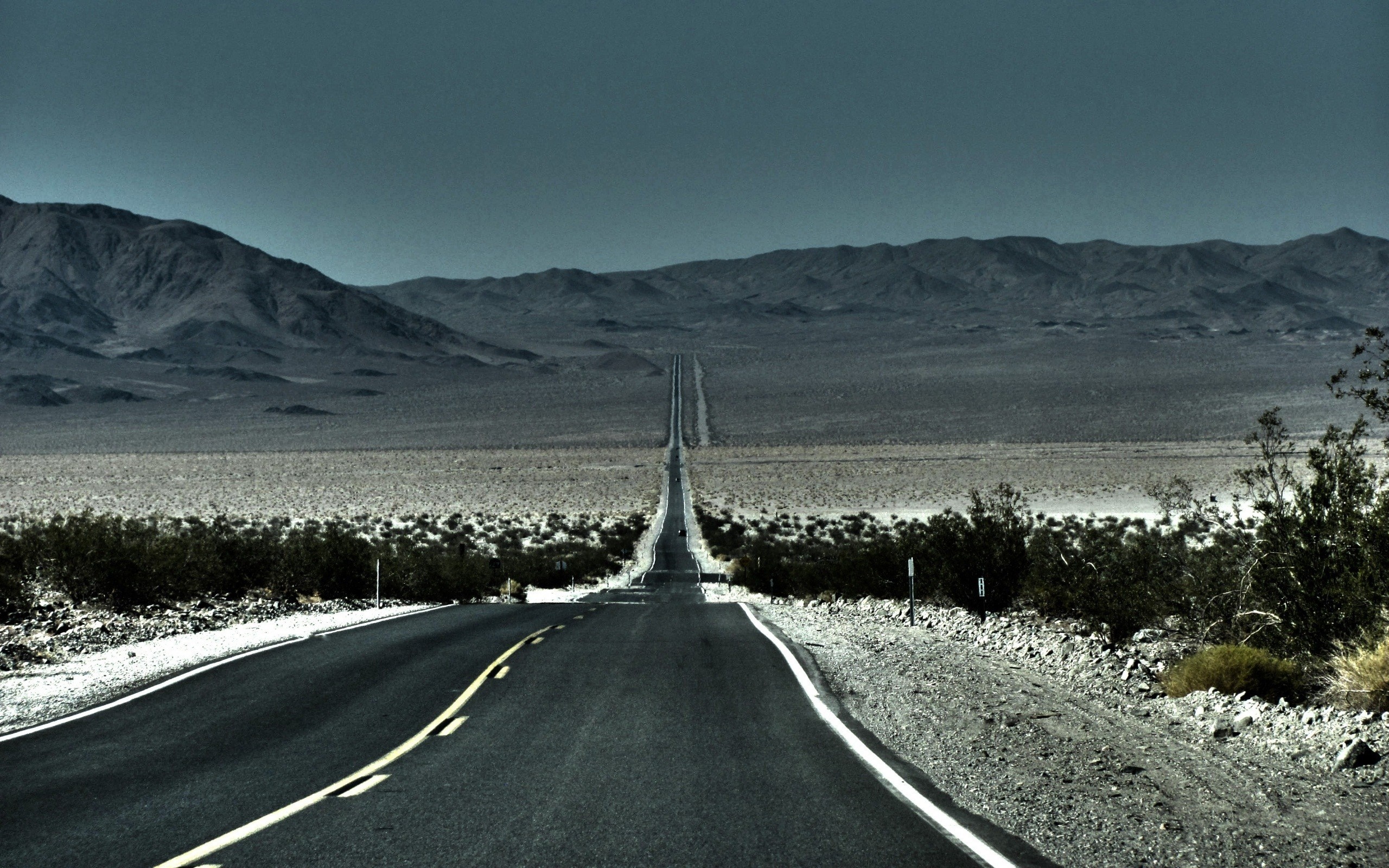 Desert highway stretching towards distant mountains under a dark sky HD Wallpaper