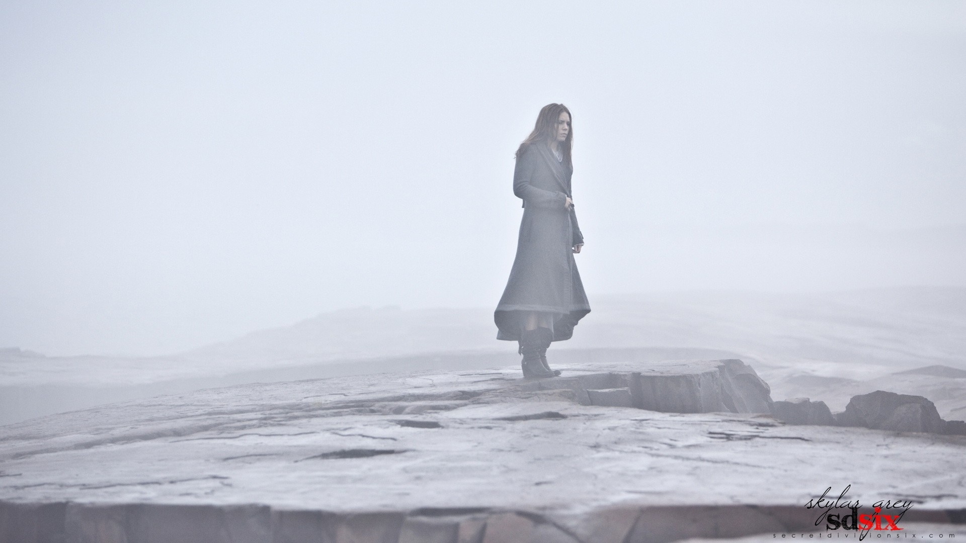 Woman in a long coat standing on a rocky outcrop in fog HD Wallpaper