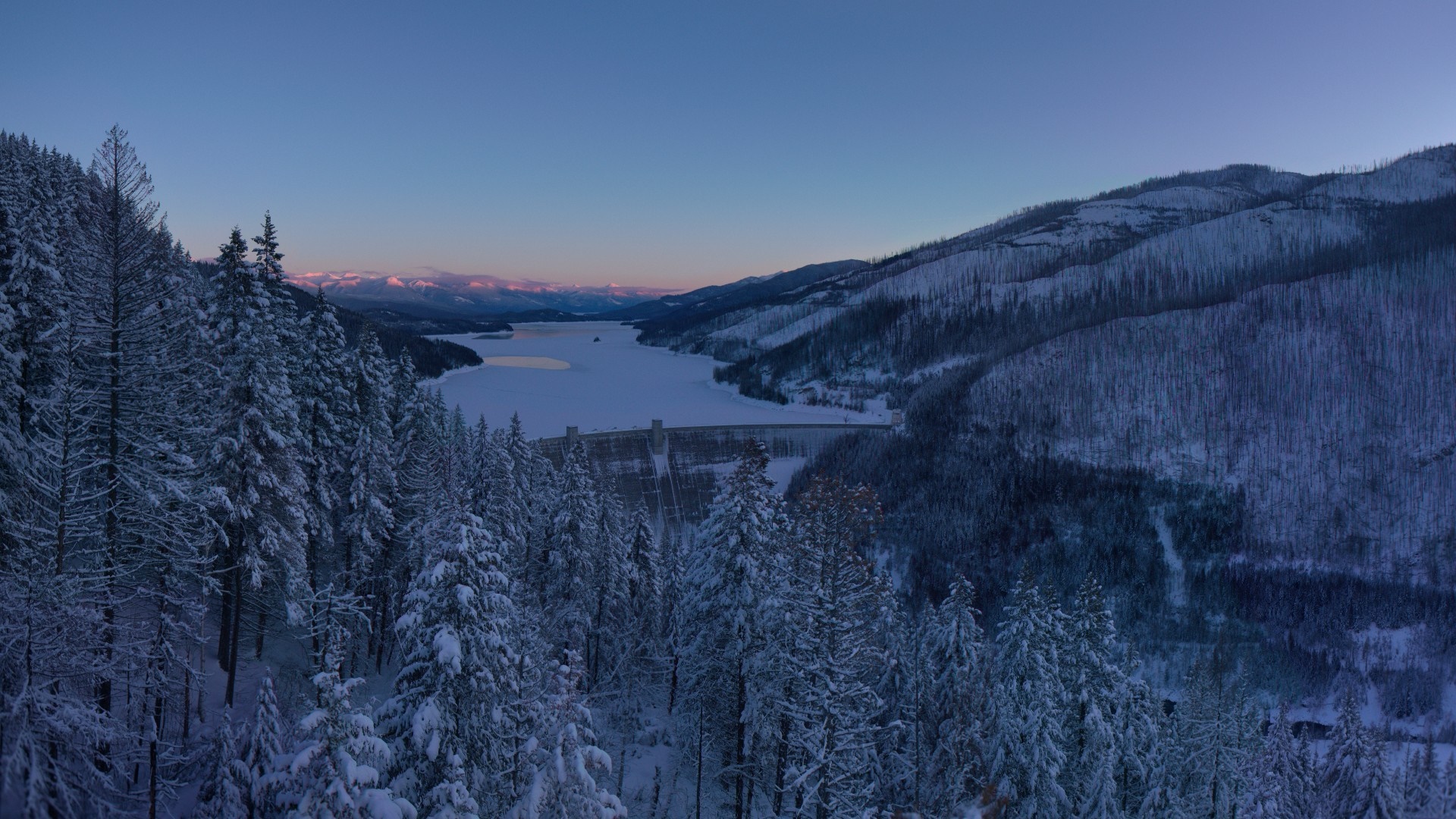 Snow-covered forest overlooking a frozen lake and dam at dawn HD Wallpaper