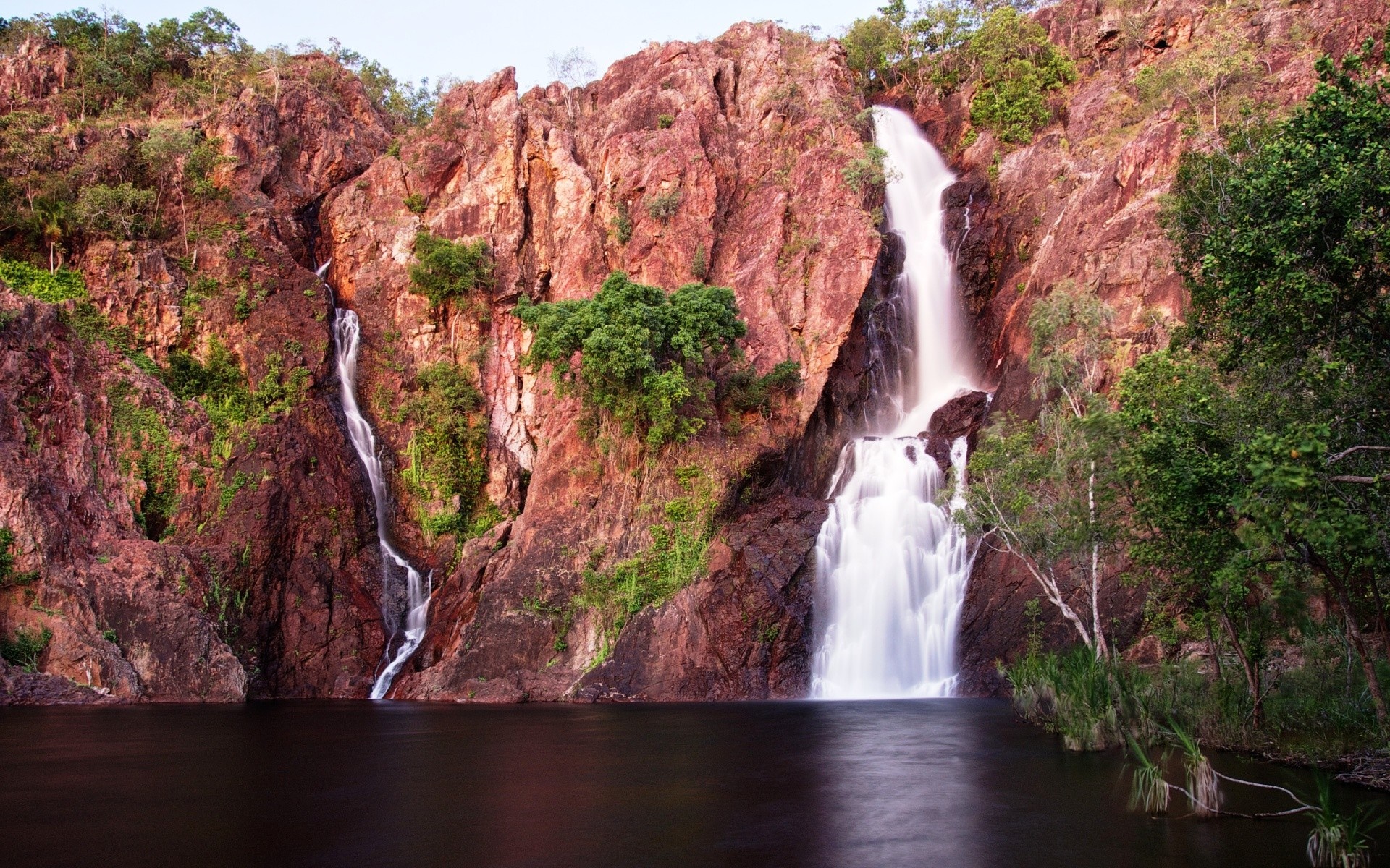 Twin waterfalls cascade down red rocky cliffs into a dark pool of water. HD Wallpaper