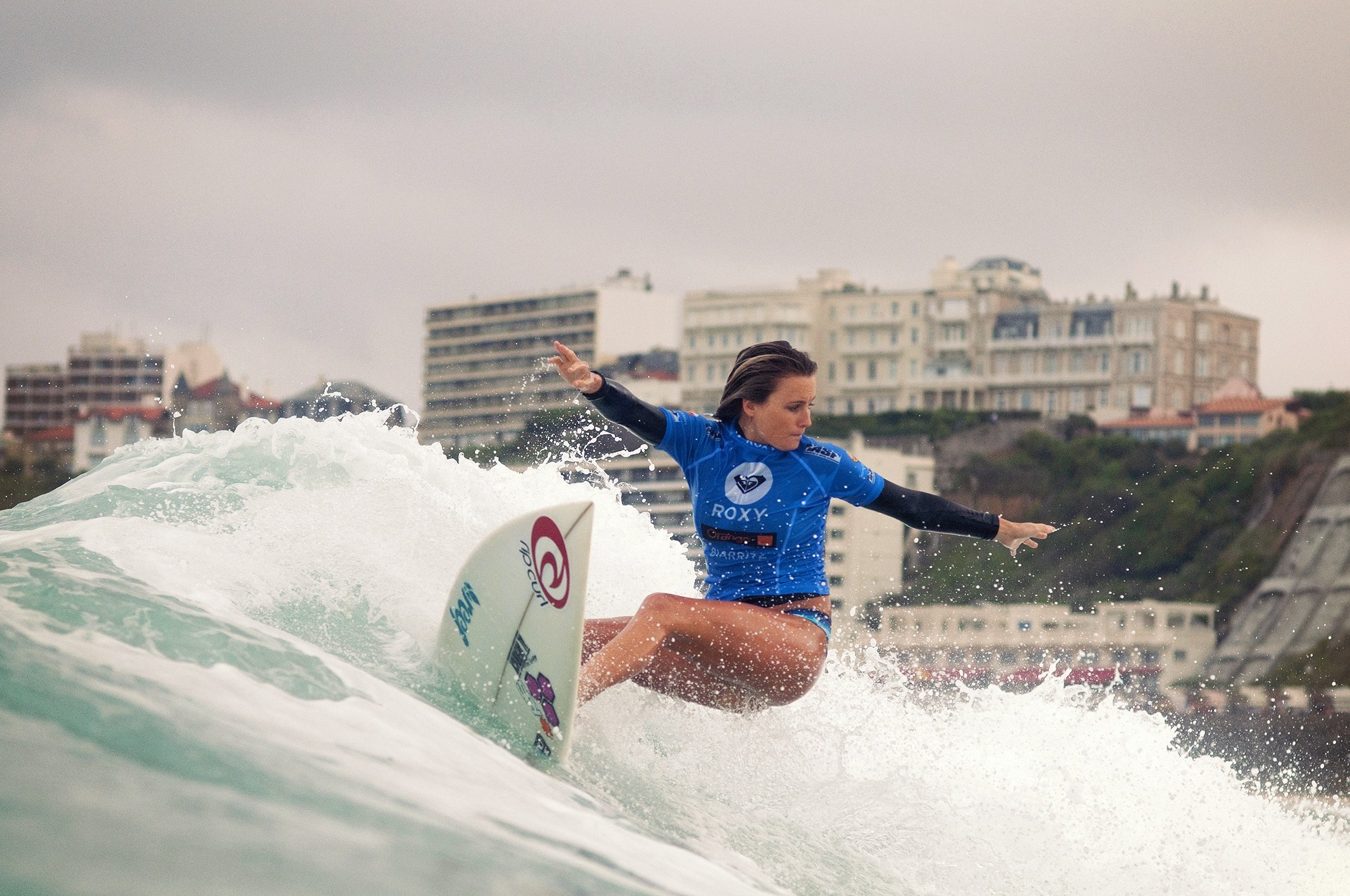 Woman surfing a wave with buildings in the background HD Wallpaper