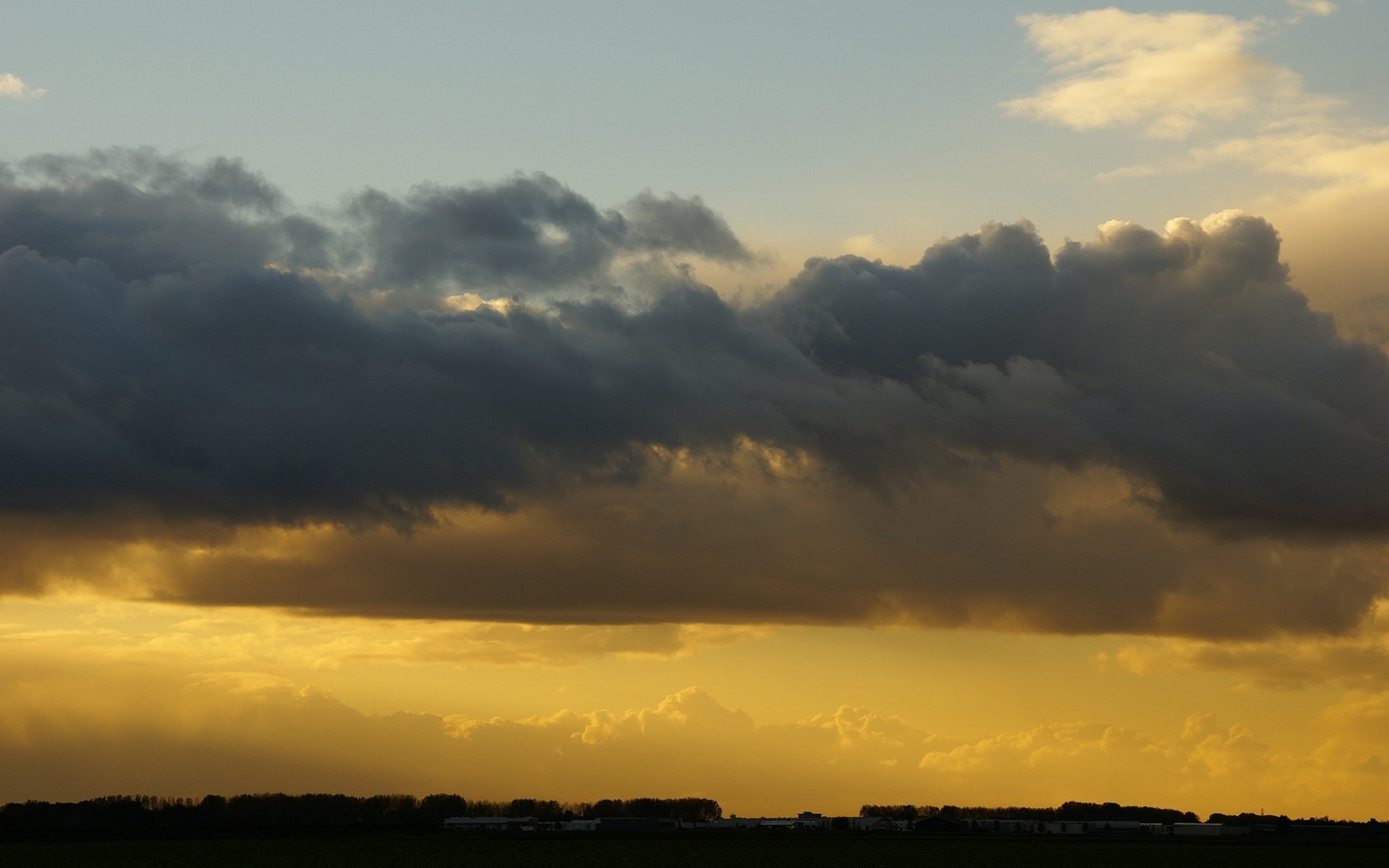 Dark clouds over a golden sunset sky above a silhouetted landscape HD Wallpaper