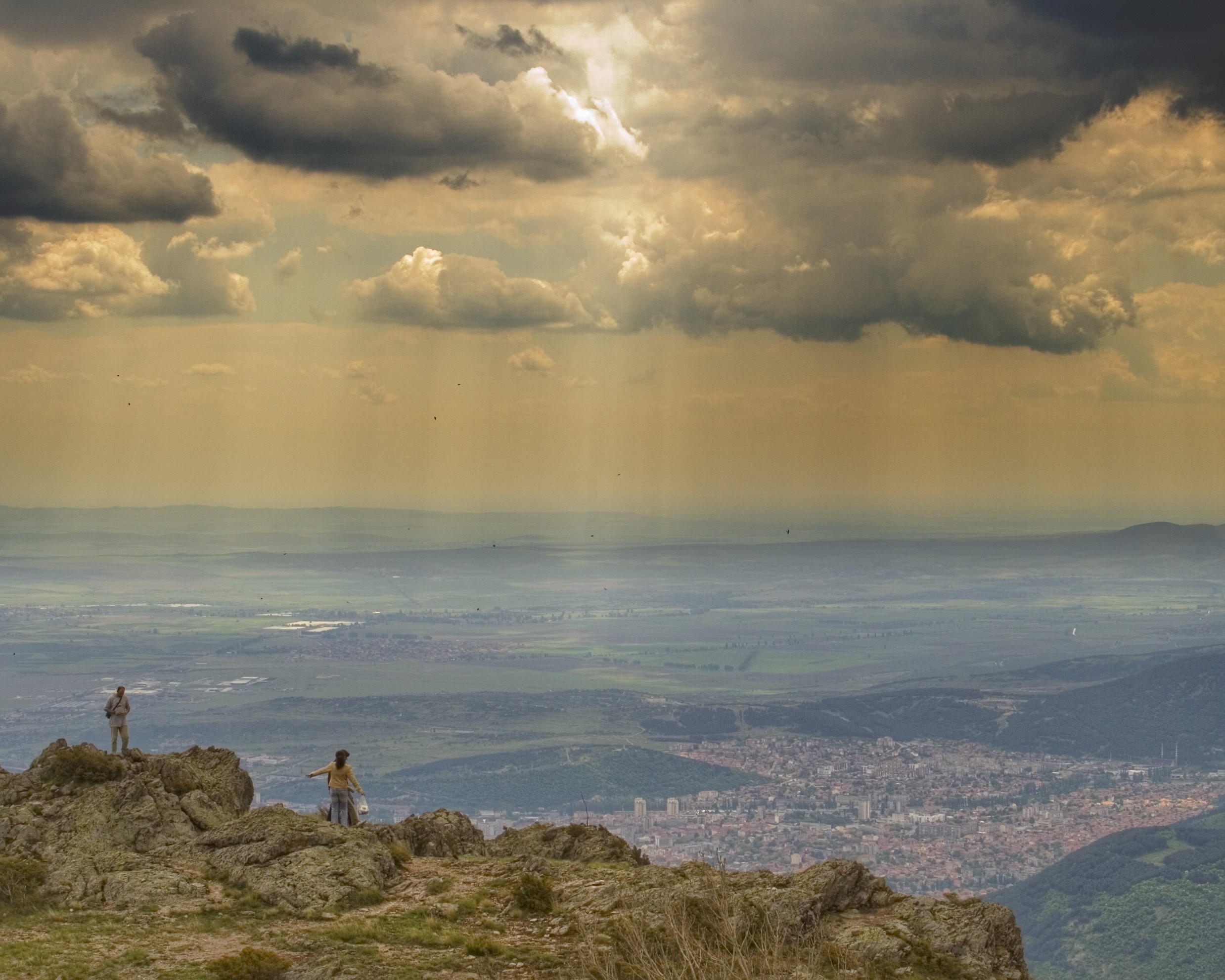 Two people on a rocky outcrop overlooking a city and valley HD Wallpaper