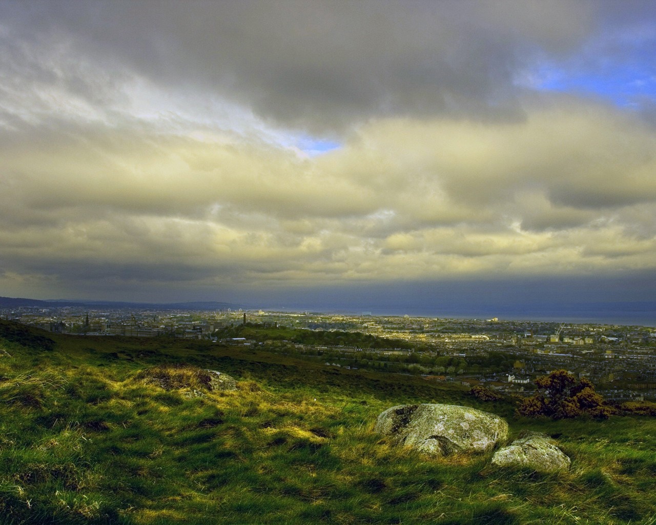 Panoramic view of a city from a grassy hill under cloudy sky HD Wallpaper