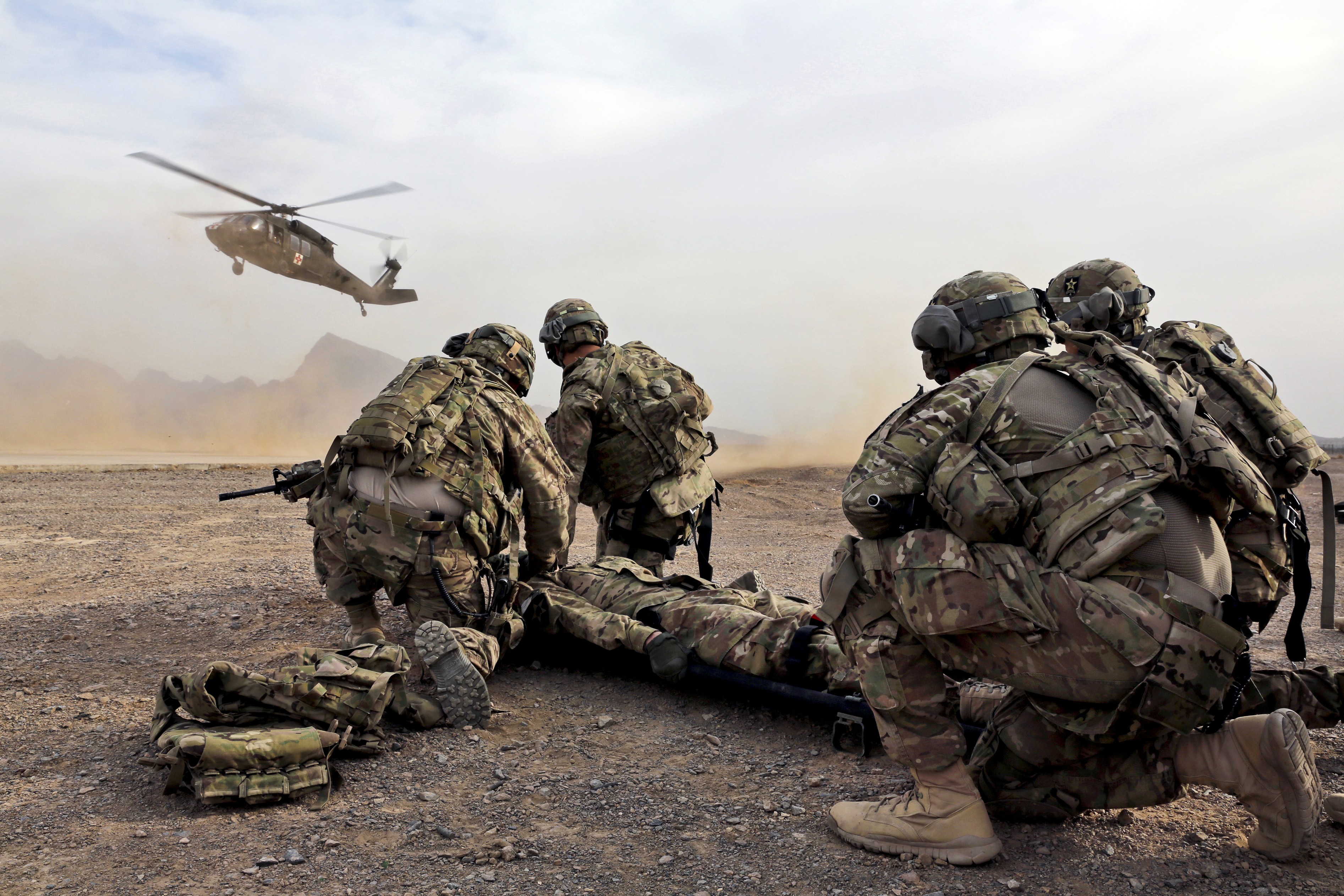 Soldiers preparing to load a casualty onto a stretcher as a helicopter approaches in a desert. HD Wallpaper