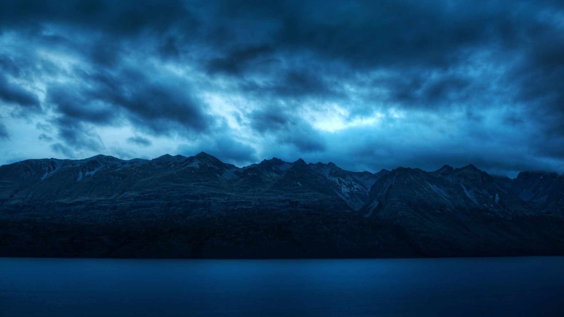 Dark Mountain Range Overlooking a Calm Lake Under Stormy Skies HD Wallpaper