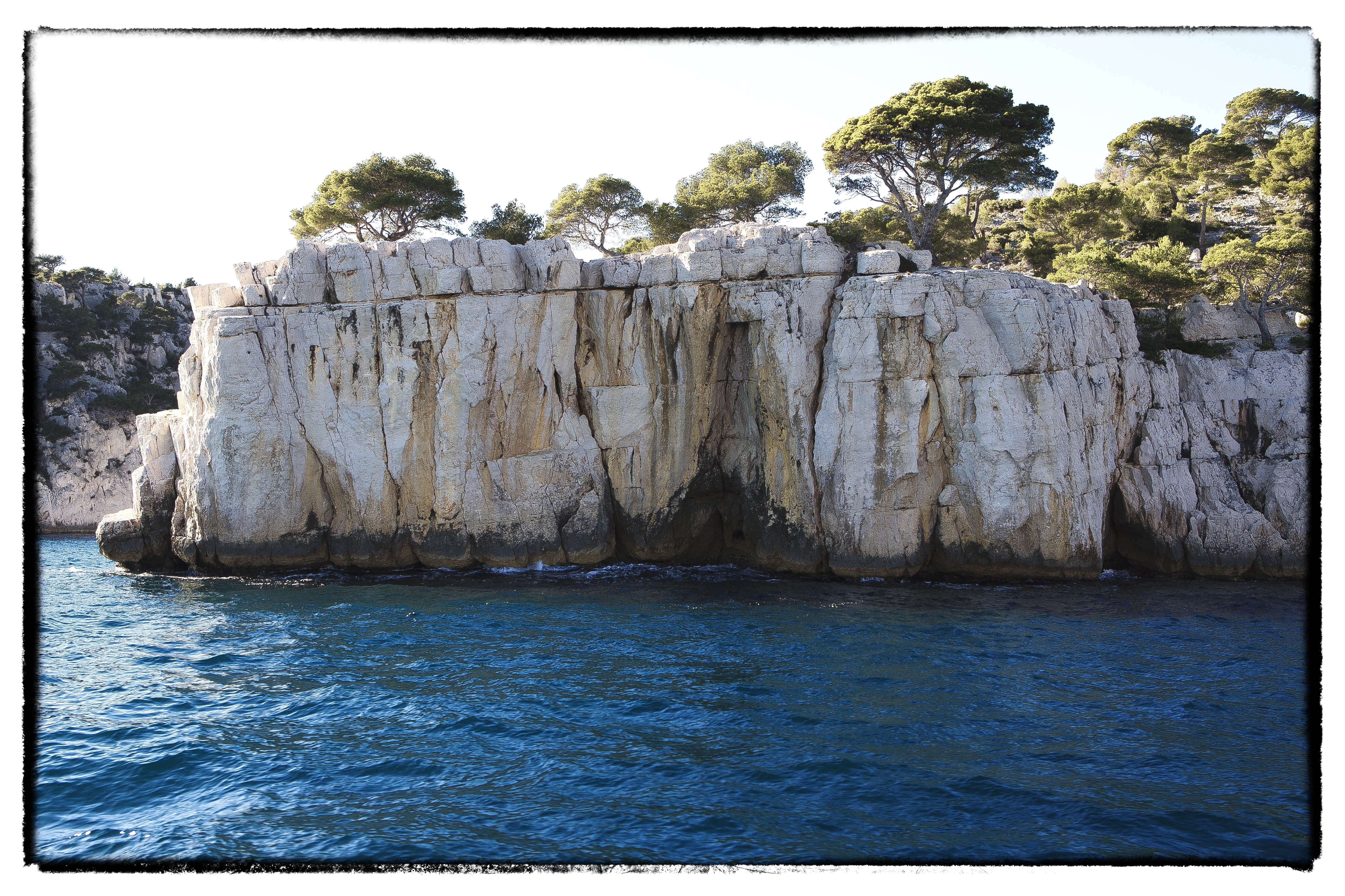 Rocky cliff face with pine trees overlooking blue sea HD Wallpaper