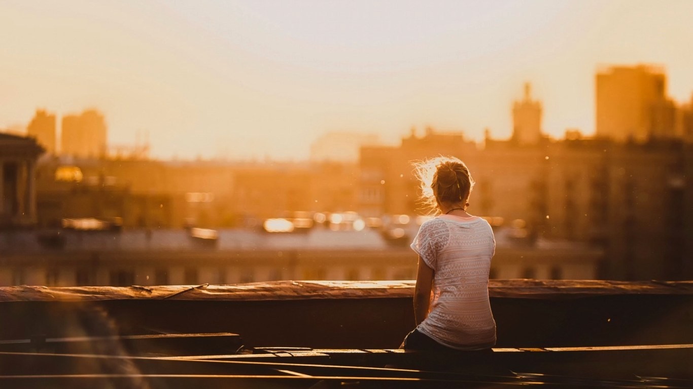 Woman sitting on rooftop at sunset overlooking city HD Wallpaper
