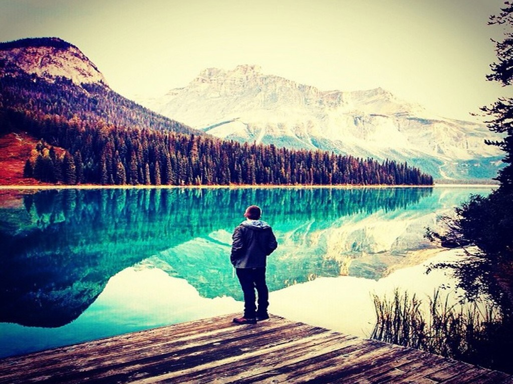 Man on wooden dock overlooking a reflective mountain lake HD Wallpaper