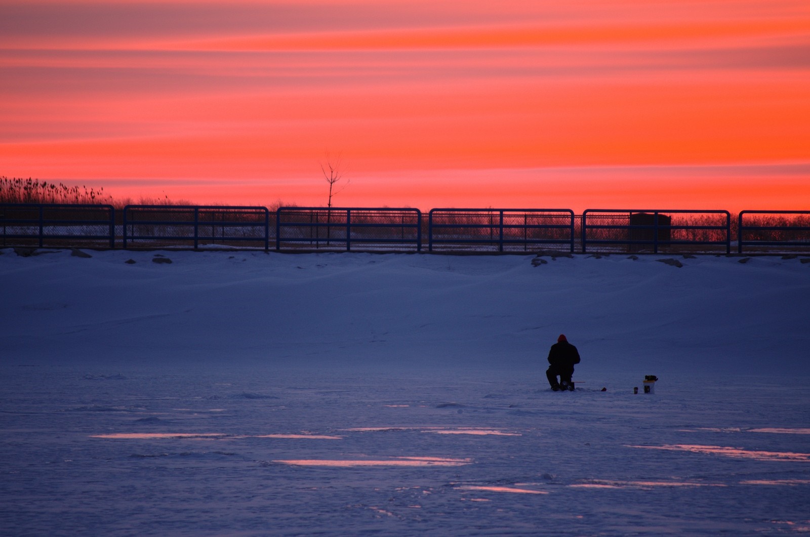 Man ice fishing during a vibrant sunset over a frozen body of water HD Wallpaper