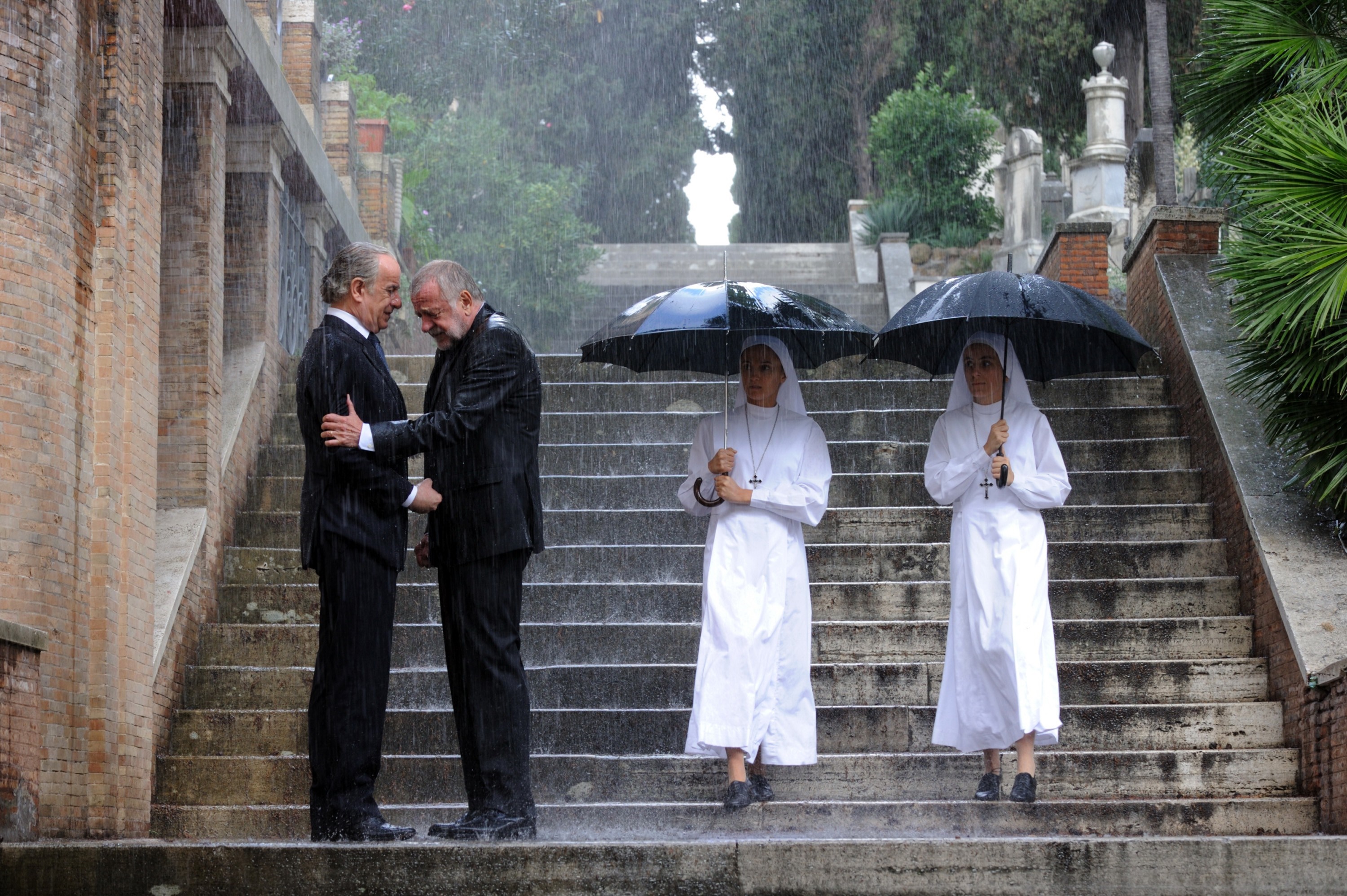 Two men in suits shake hands in the rain with nuns holding umbrellas HD Wallpaper