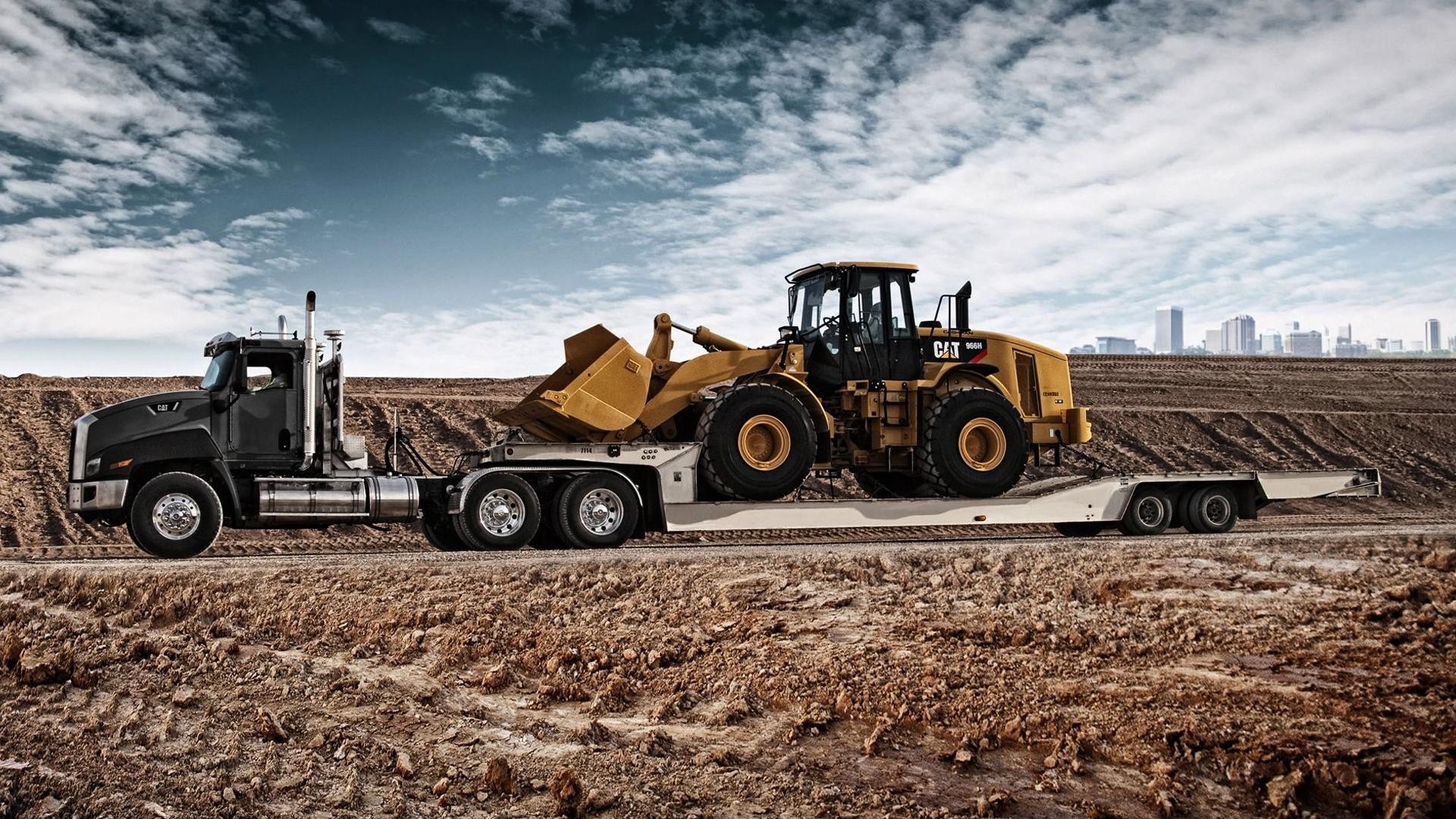 Black semi-truck hauling a yellow CAT wheel loader on a trailer HD Wallpaper