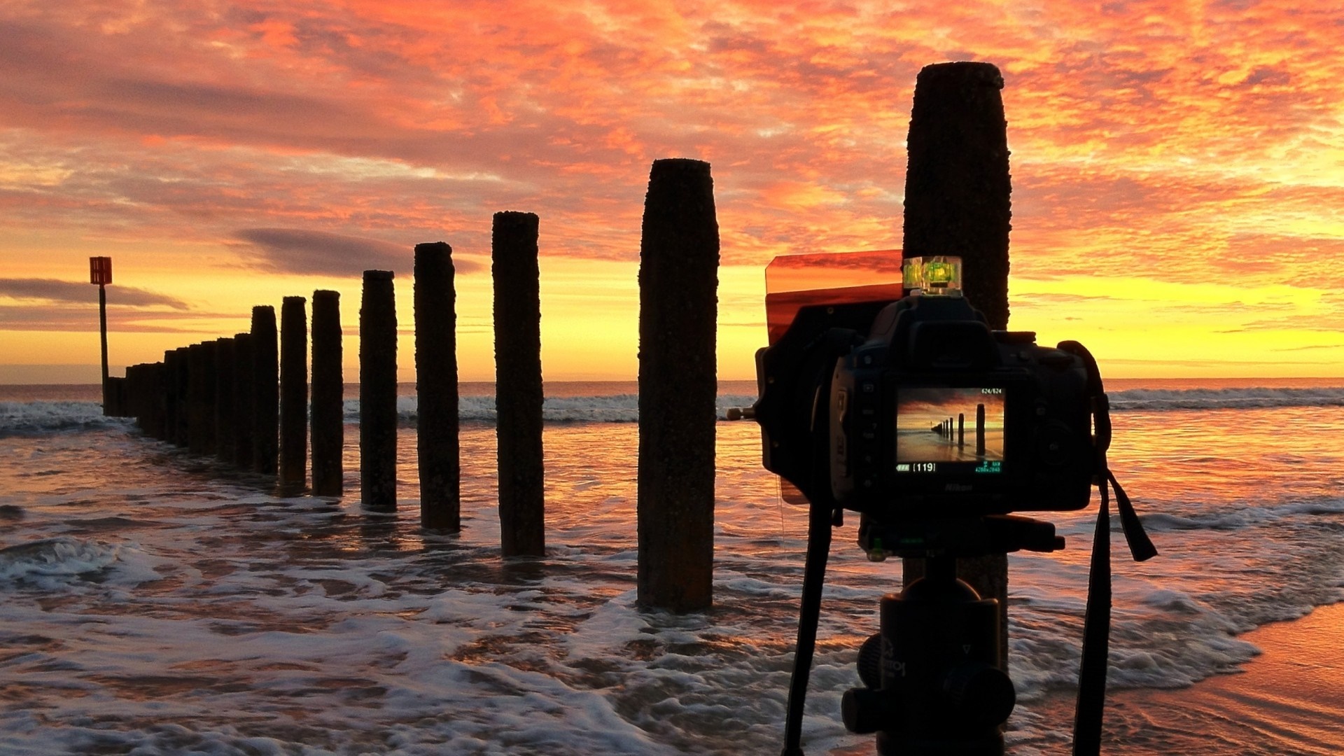 Camera photographing a sunset over the sea with wooden posts HD Wallpaper