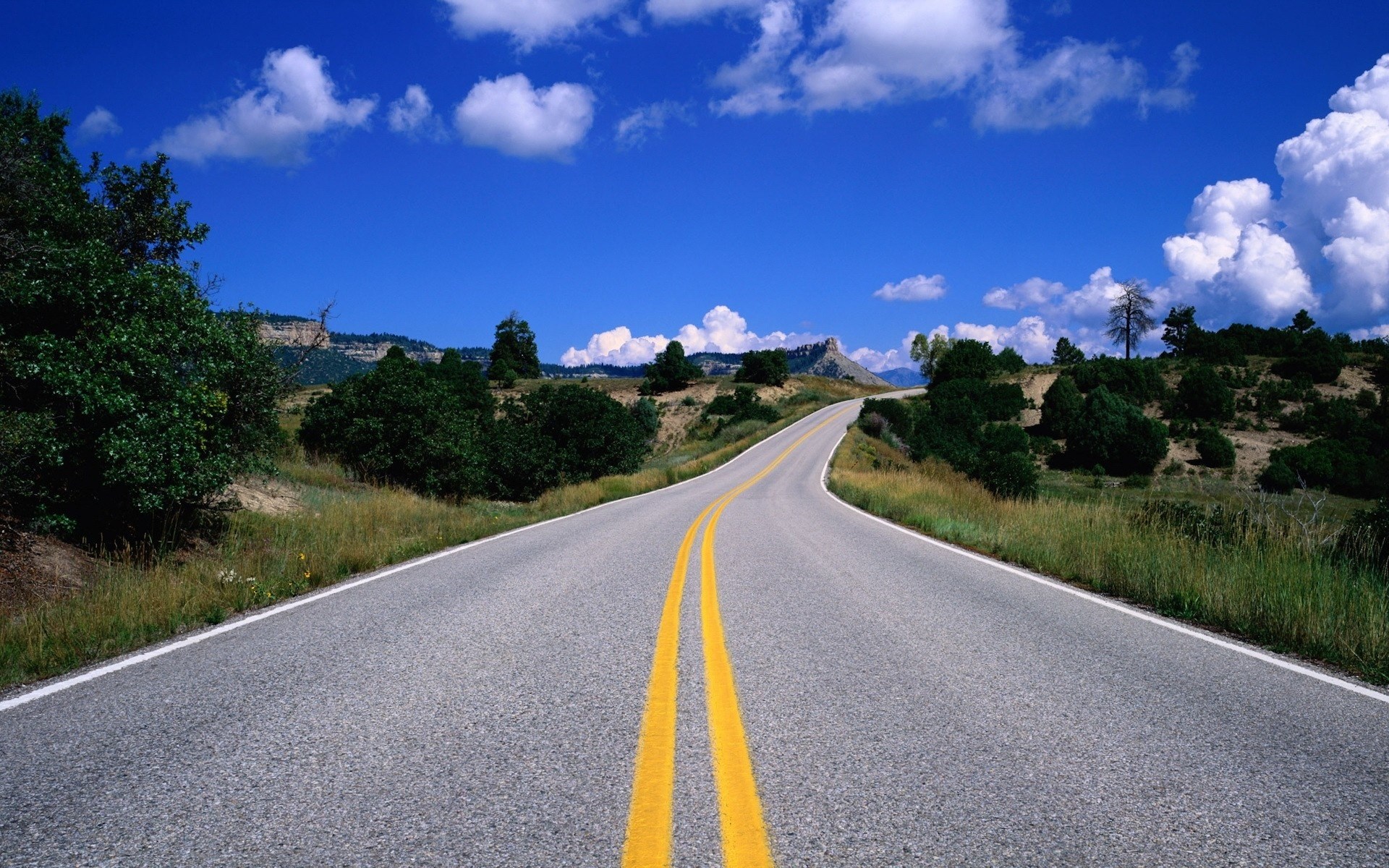 Empty road winding through a hilly landscape under a blue sky HD Wallpaper