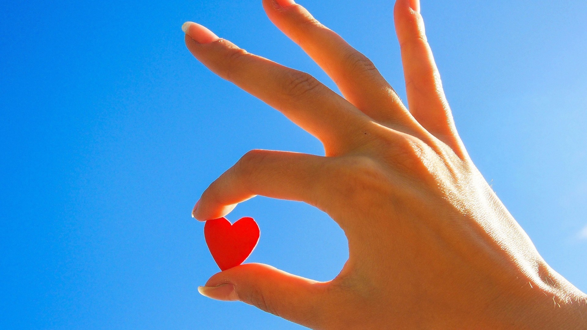 Hand holding a small red heart against a blue sky HD Wallpaper