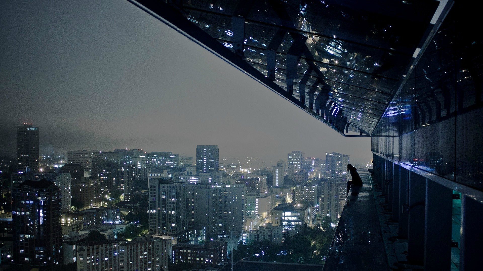 Person sitting on edge of skyscraper overlooking city at night HD Wallpaper