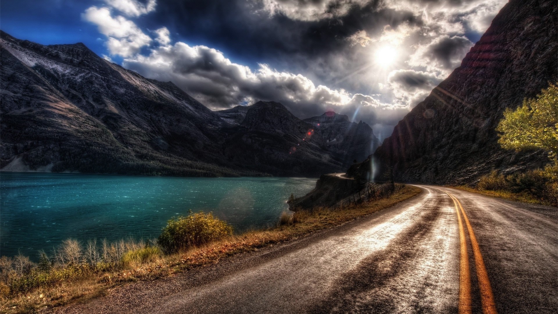 Road alongside a lake and mountains under dramatic sunlight HD Wallpaper