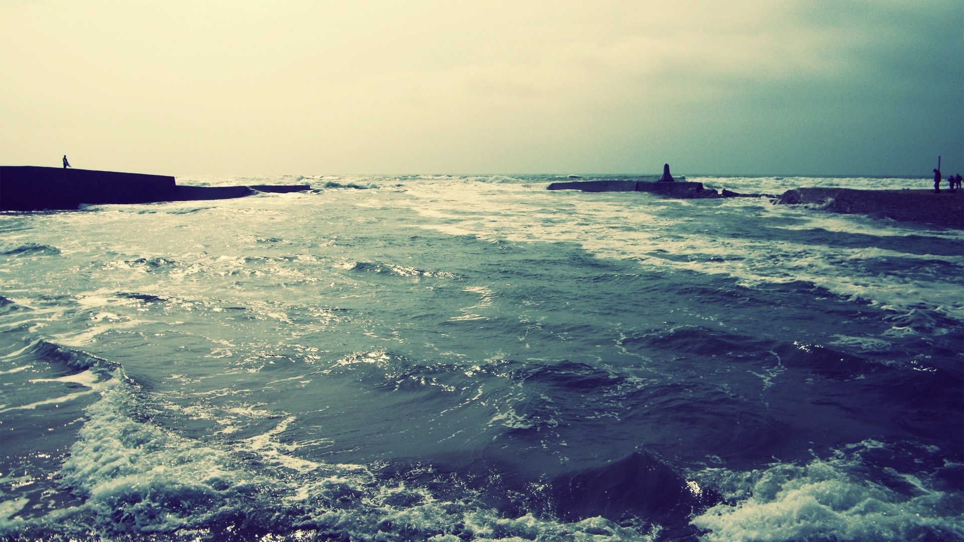 Waves crashing on a pier with people in the distance HD Wallpaper