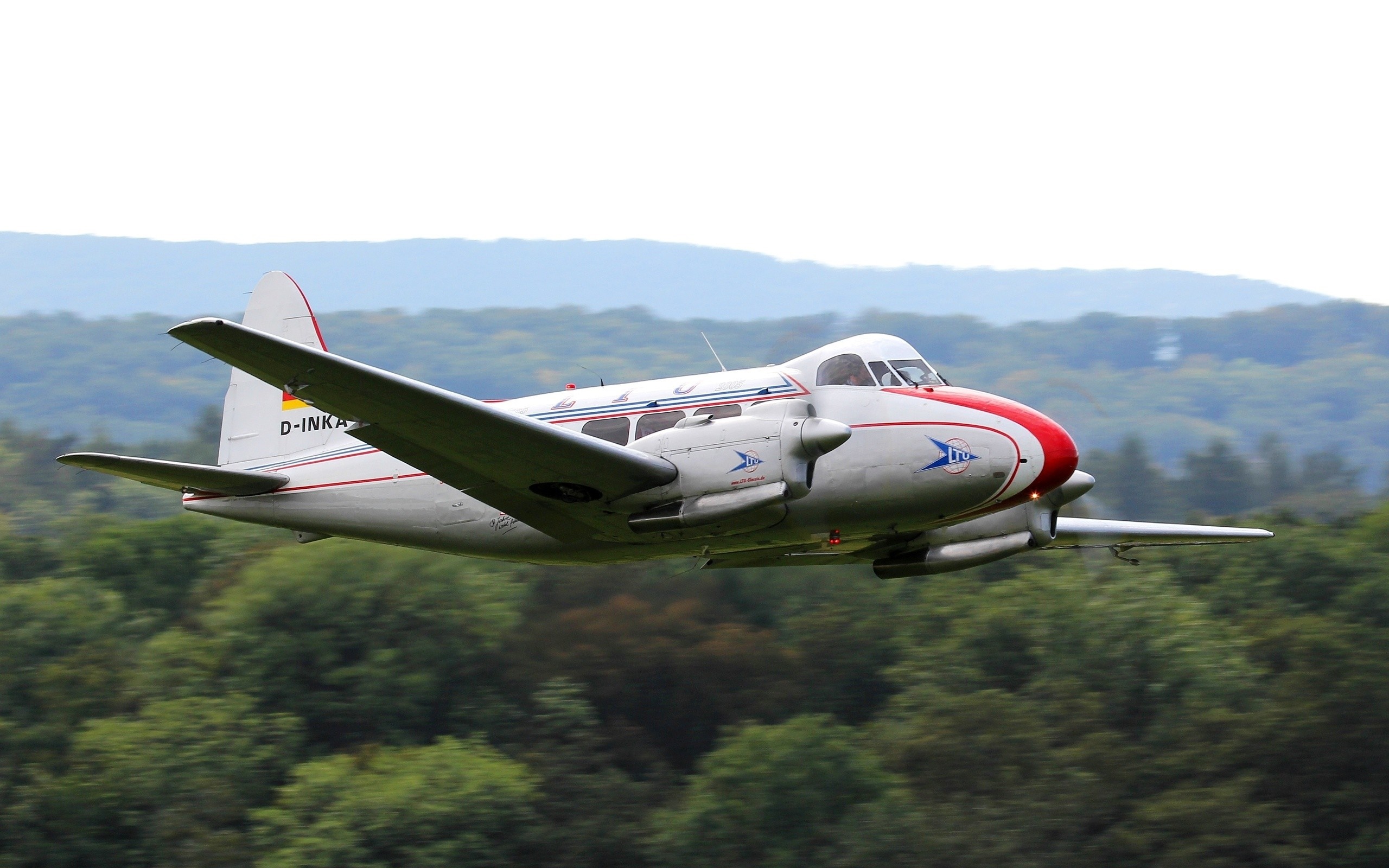 A white twin-engine airplane with red accents flying over trees HD Wallpaper