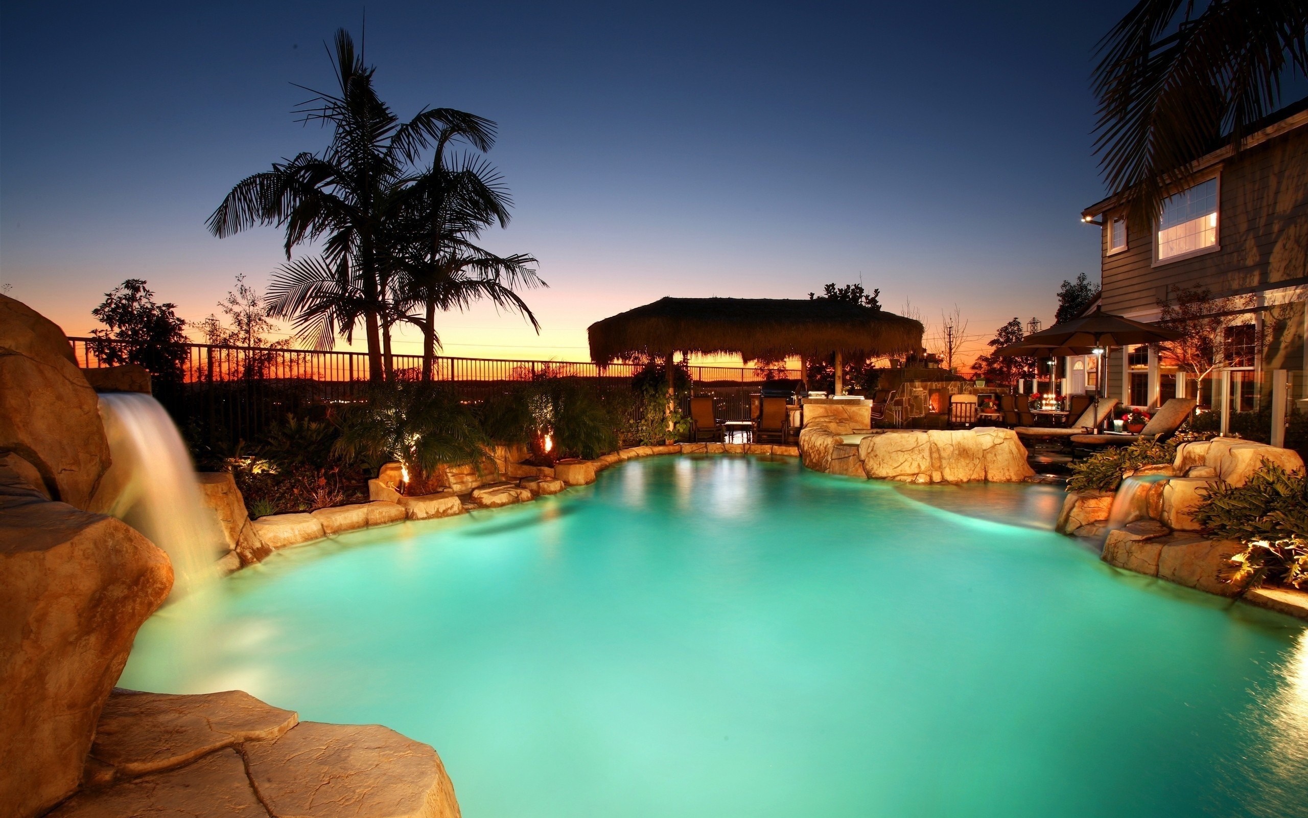 Luxury swimming pool at dusk with waterfall and tiki bar HD Wallpaper