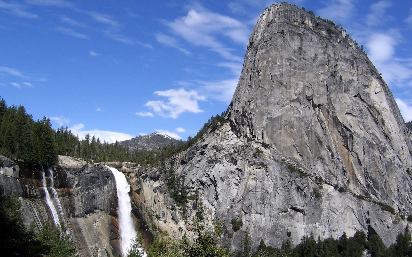 Waterfall cascading down a rocky cliff face with a large granite dome mountain in the background under a blue sky with clouds. HD Wallpaper