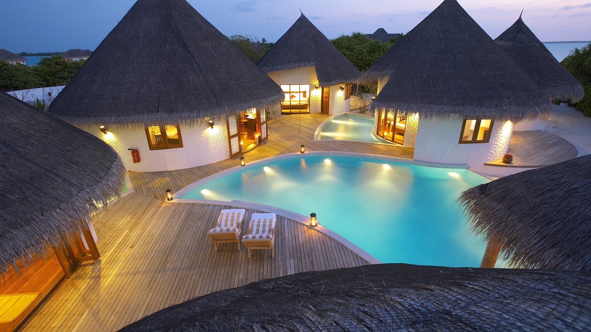 Luxury resort with thatched-roof bungalows and swimming pool at dusk HD Wallpaper