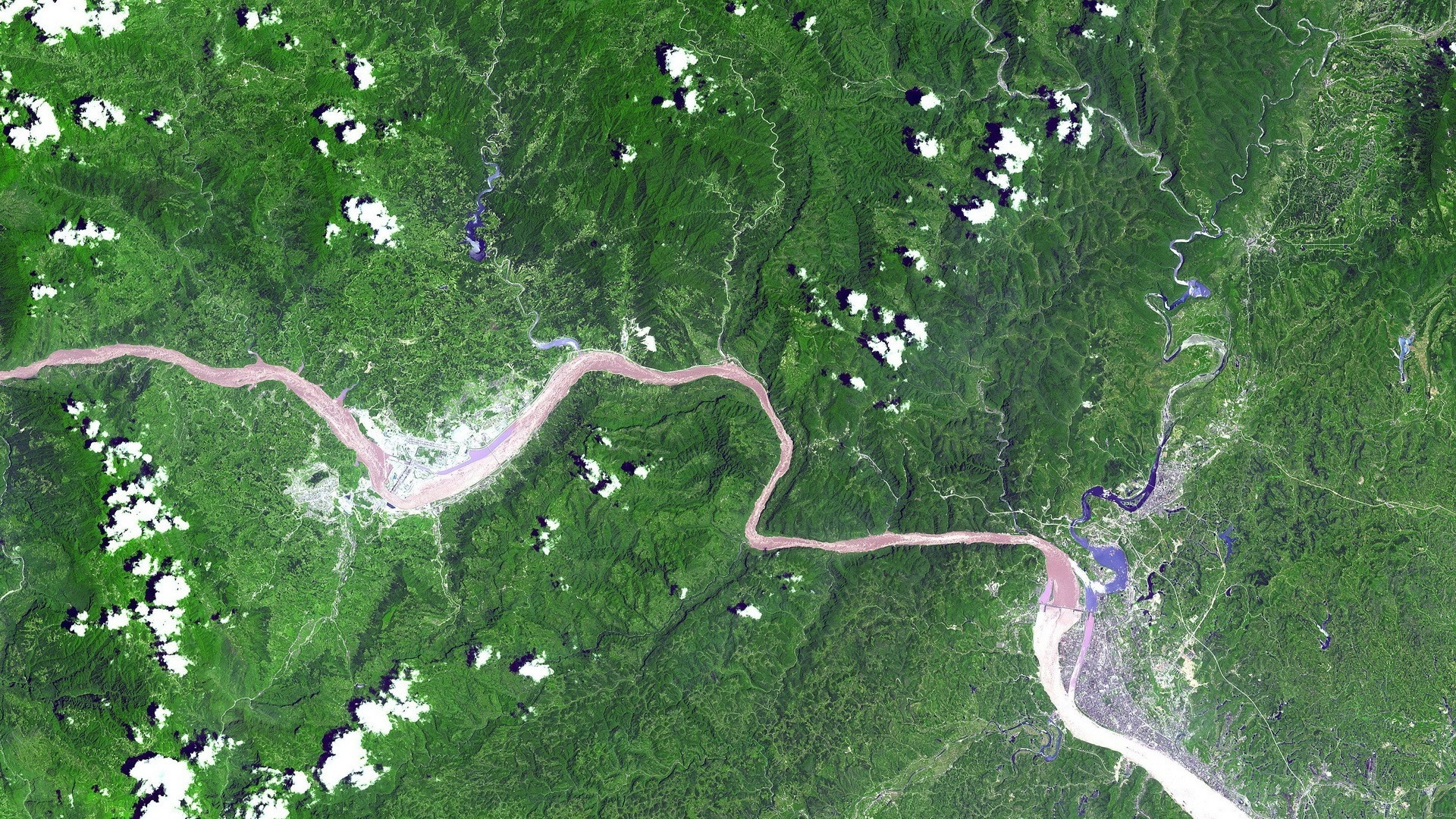 Aerial view of a wide, muddy river flowing through a dense green forest with scattered clouds. HD Wallpaper
