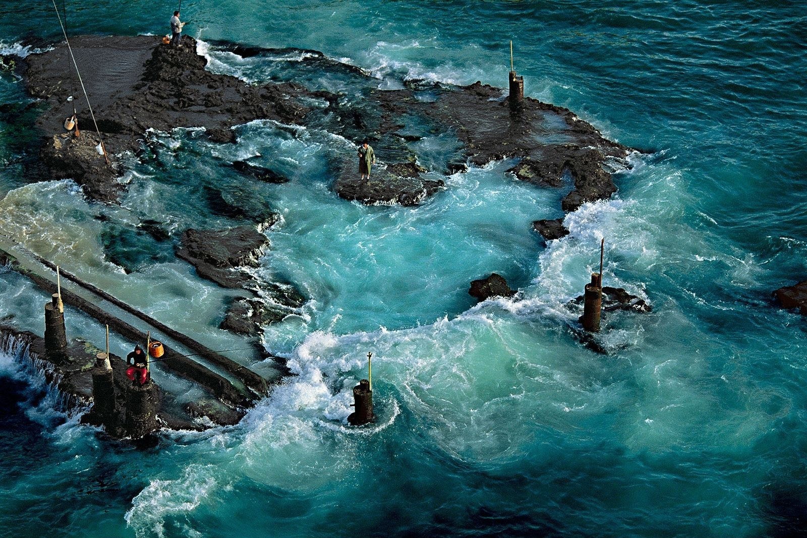 People fishing on rocky outcrops in rough turquoise ocean waves HD Wallpaper