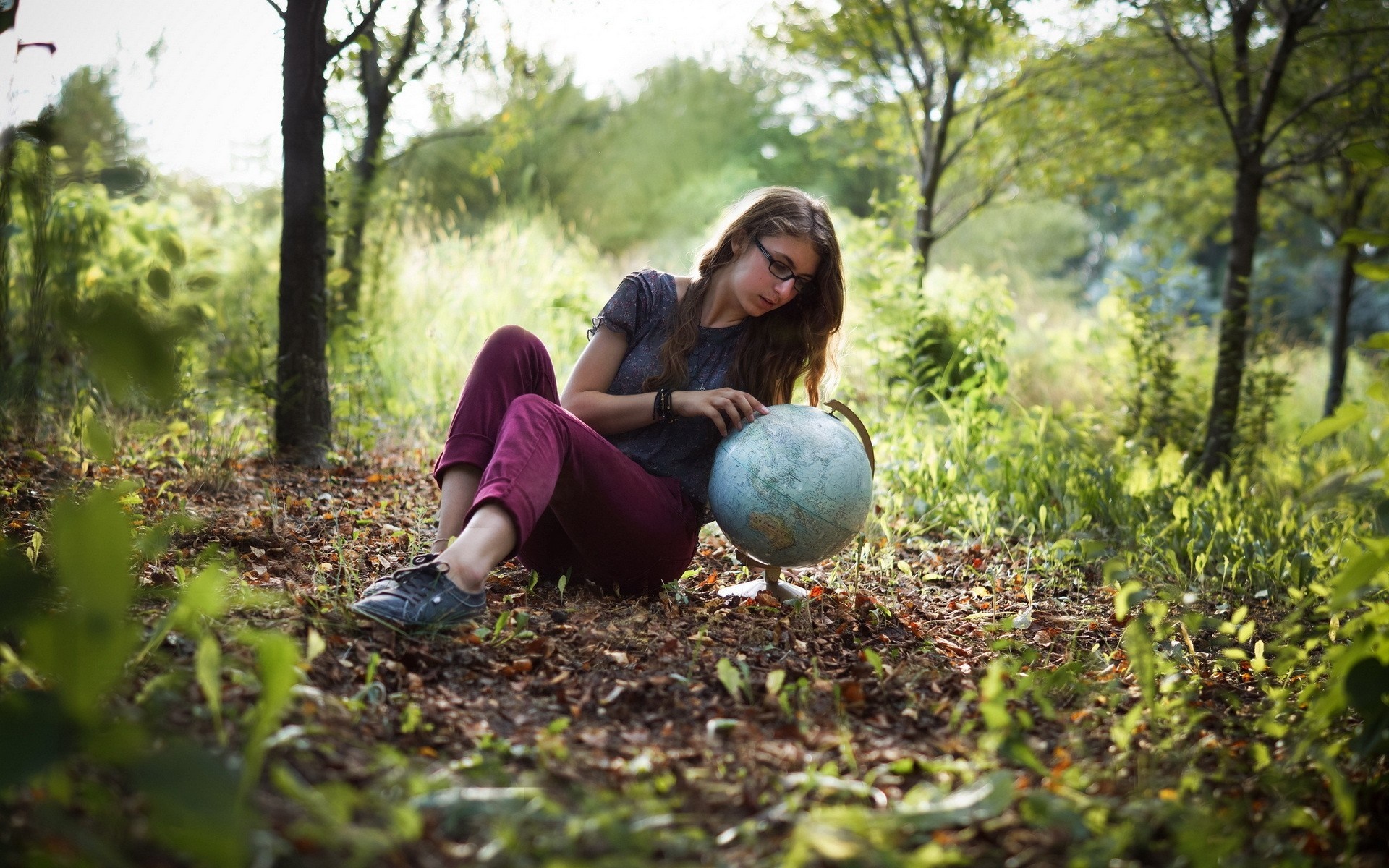 Young woman with glasses looking at a globe in a forest HD Wallpaper