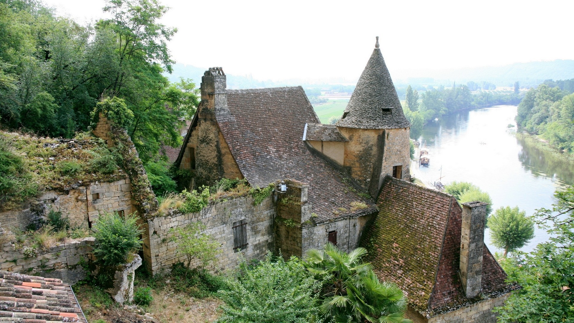 Overgrown stone buildings on a hillside overlooking a river HD Wallpaper