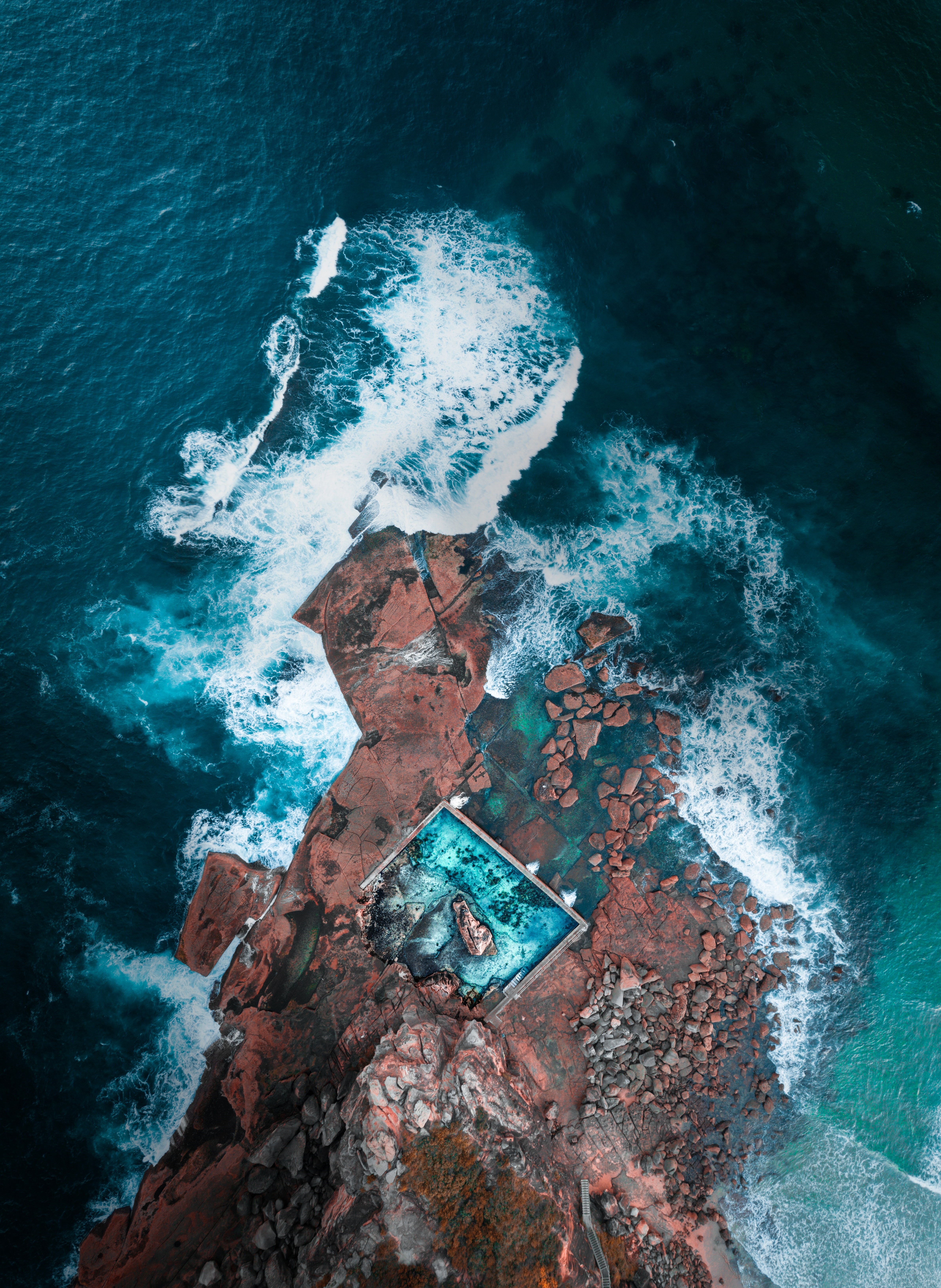 Aerial view of ocean waves crashing on rocky coastline with swimming pool HD Phone Wallpaper