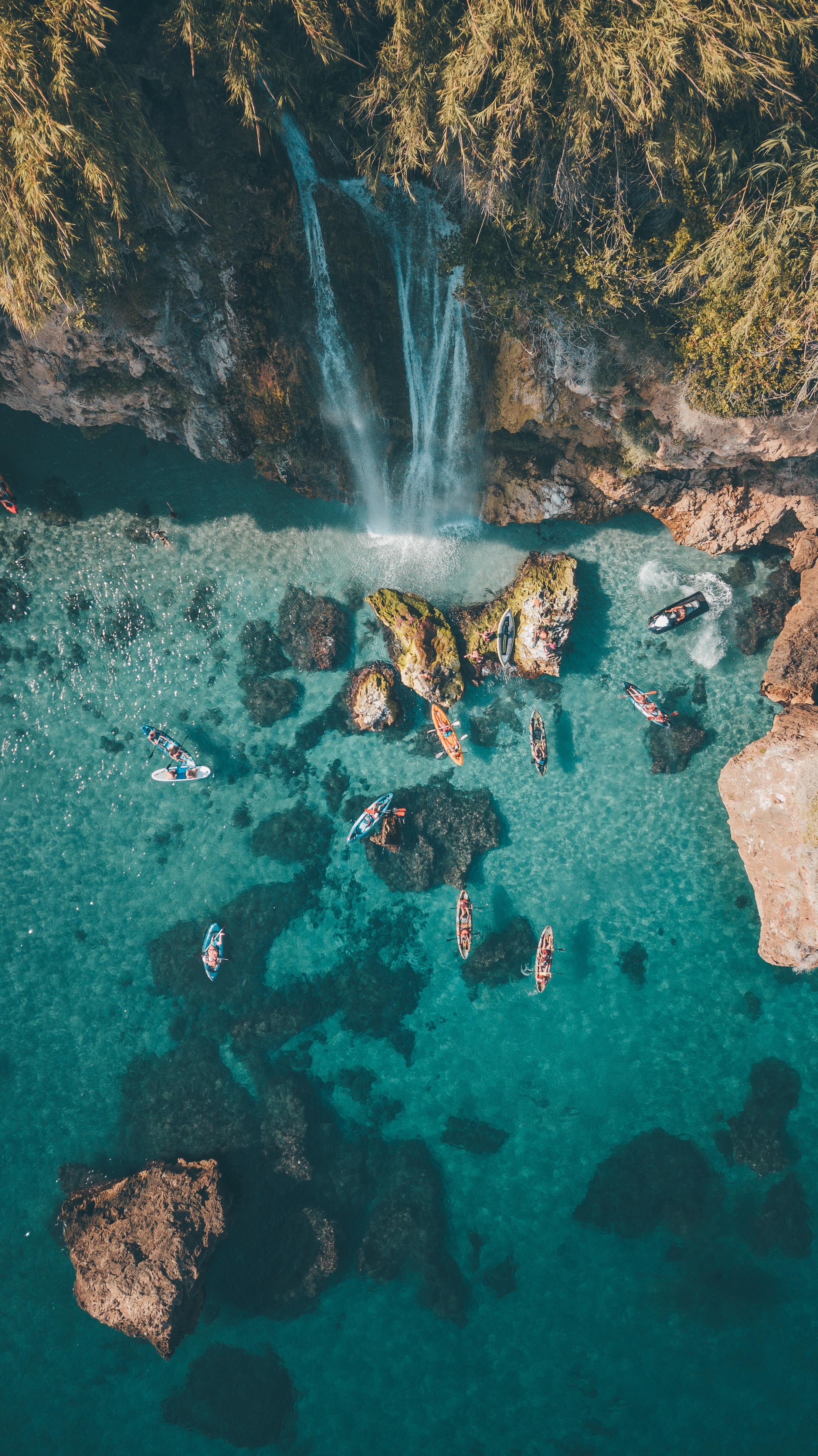 Aerial view of a waterfall cascading into the ocean with kayaks and boats HD Phone Wallpaper