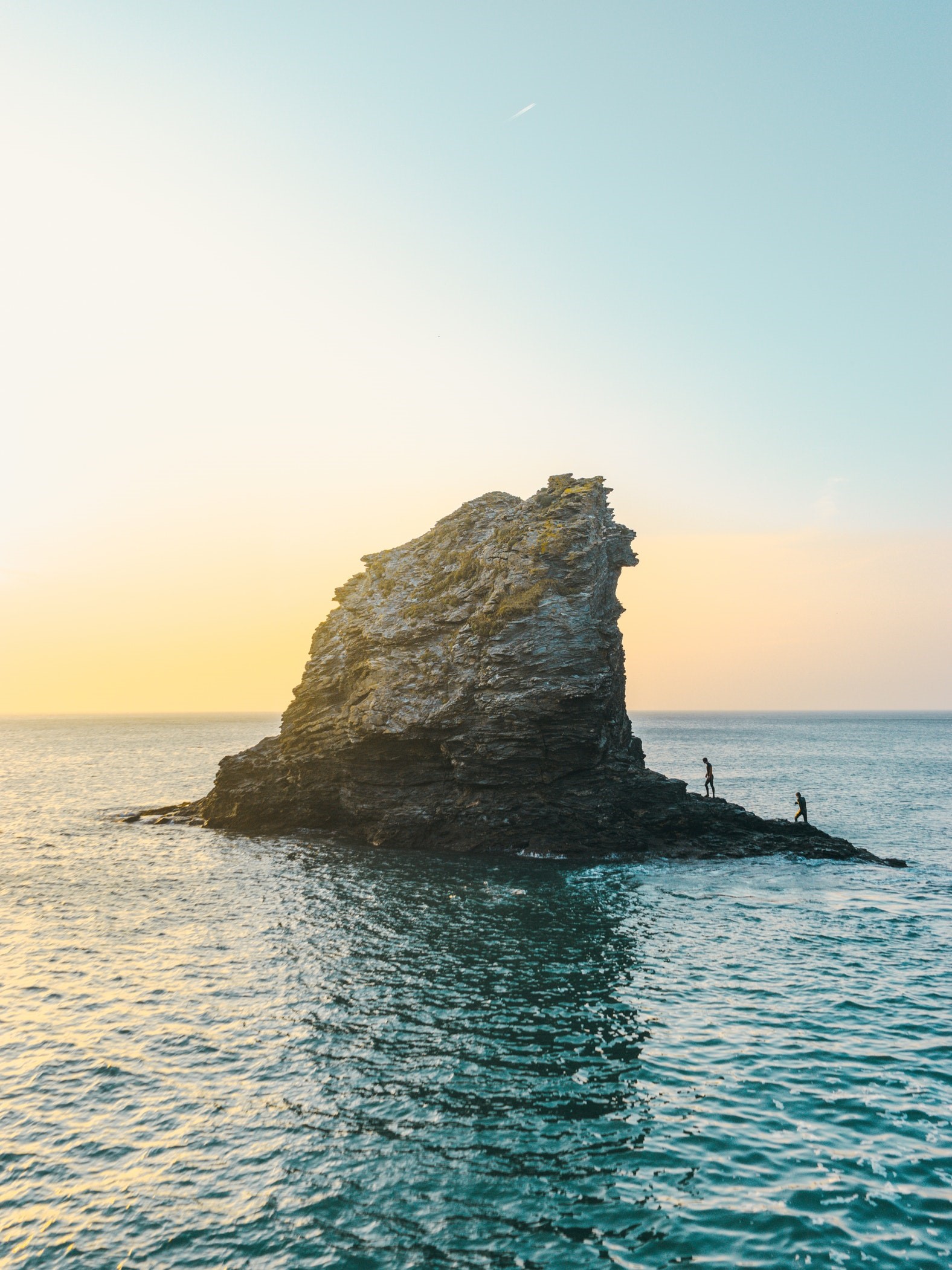 Two people on a rock formation in the ocean at sunset HD Phone Wallpaper