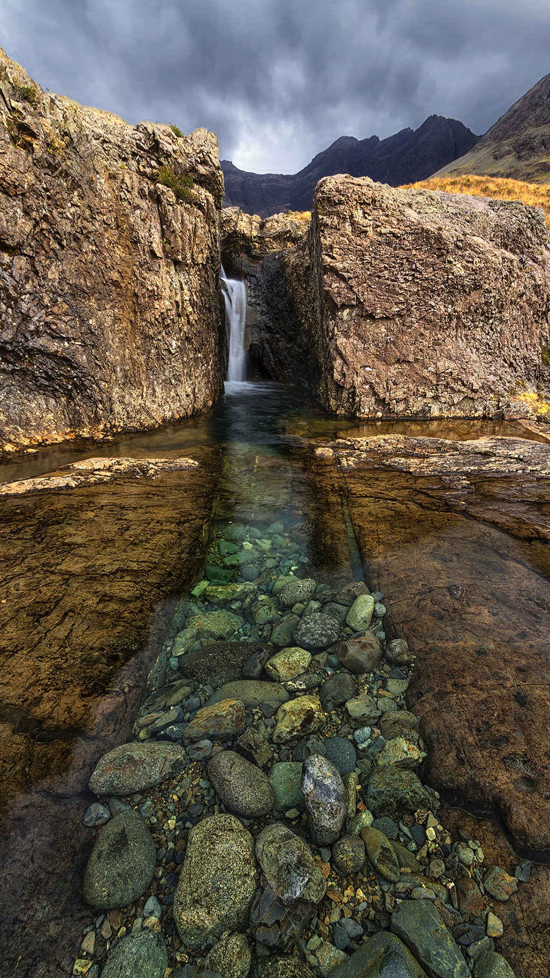 Waterfall flowing between rocks with mountains in the background HD Phone Wallpaper