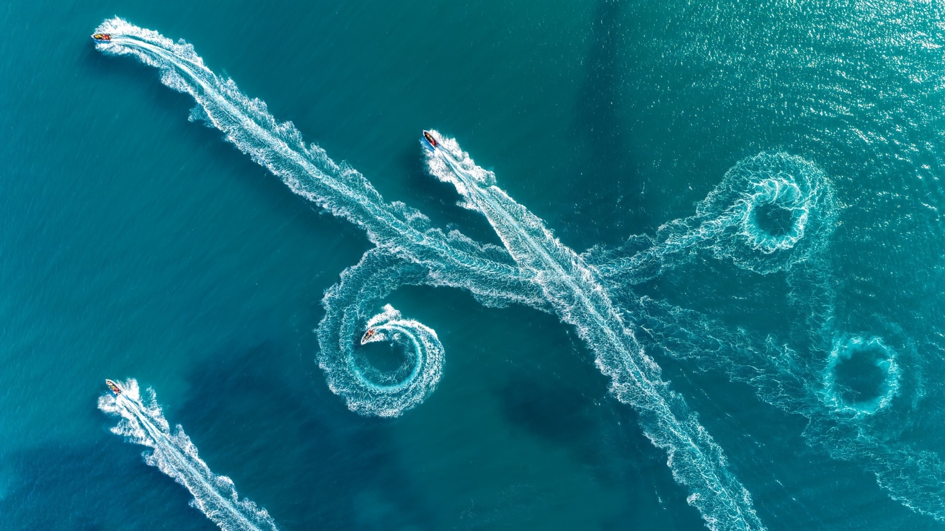 Jet skis creating swirling water trails from above HD Wallpaper