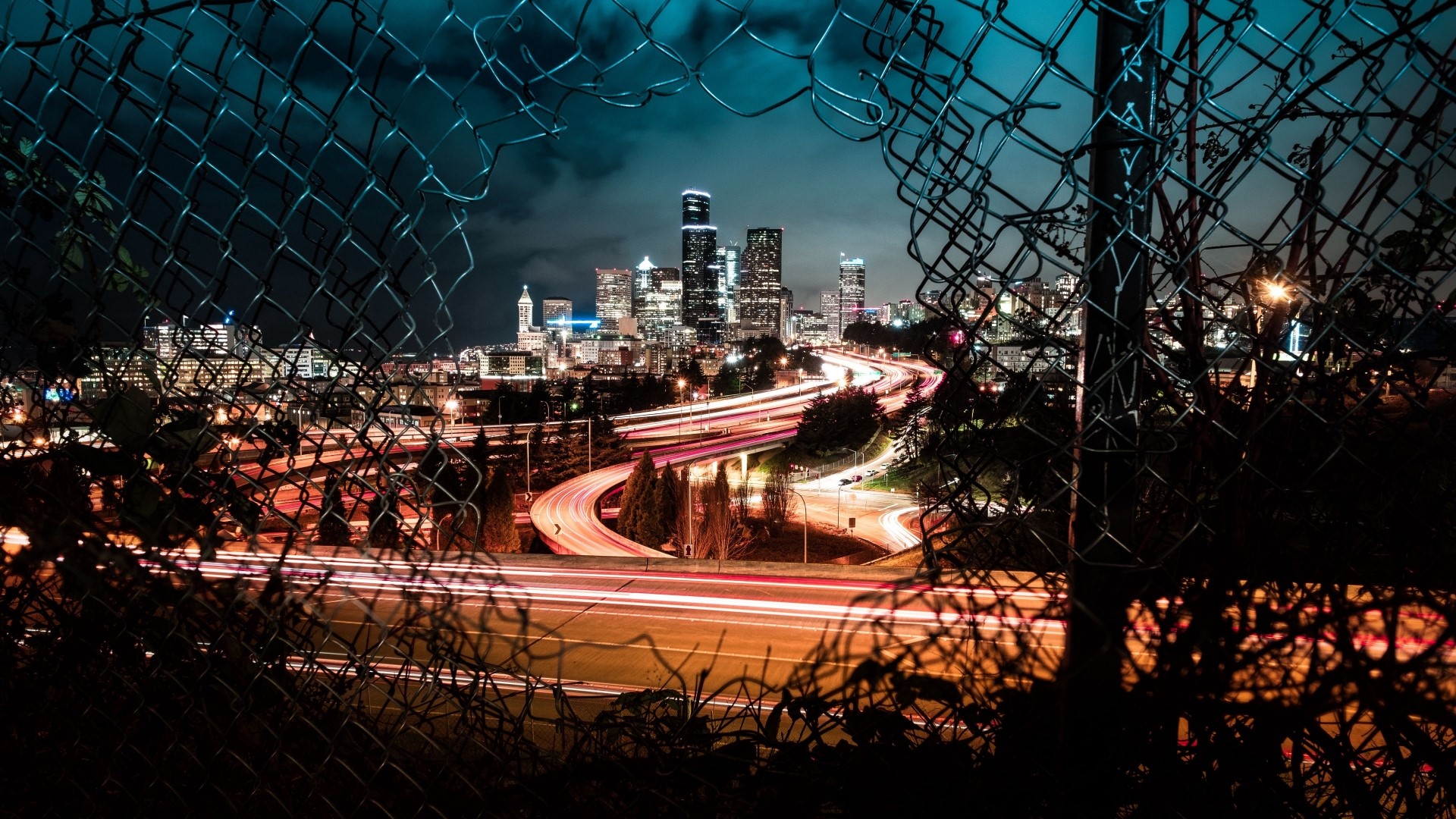 Night city skyline with light trails through a chain-link fence HD Wallpaper