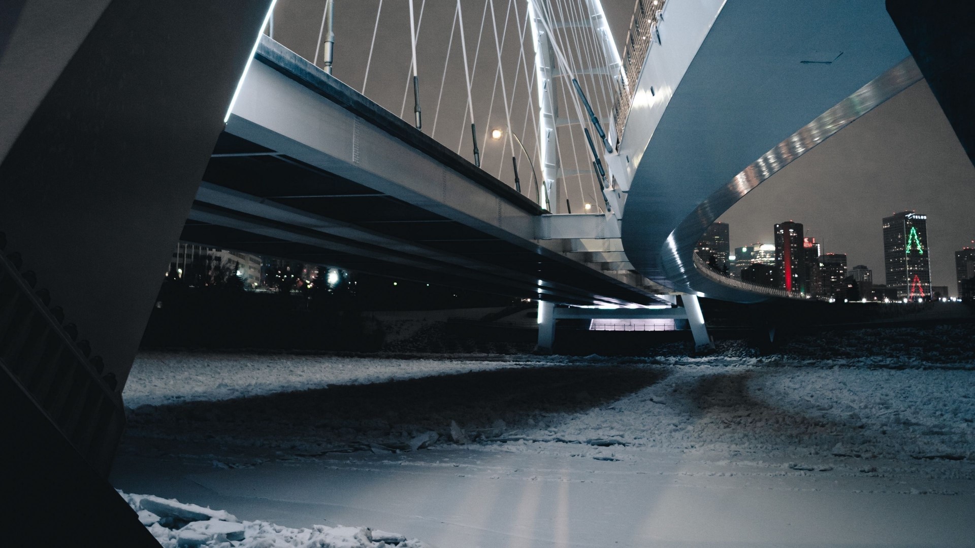 Bridge over frozen river at night with city skyline HD Wallpaper