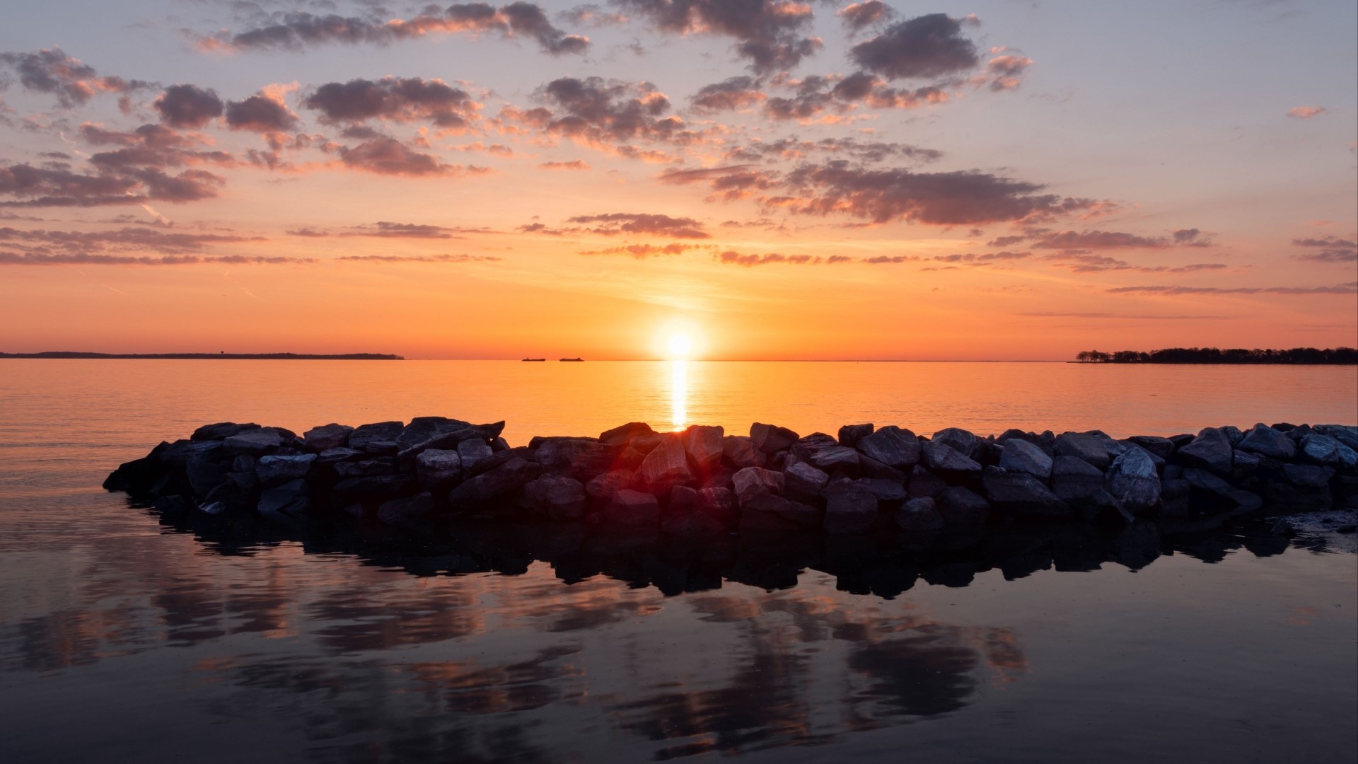 Sunset over a calm body of water with a rock jetty HD Wallpaper