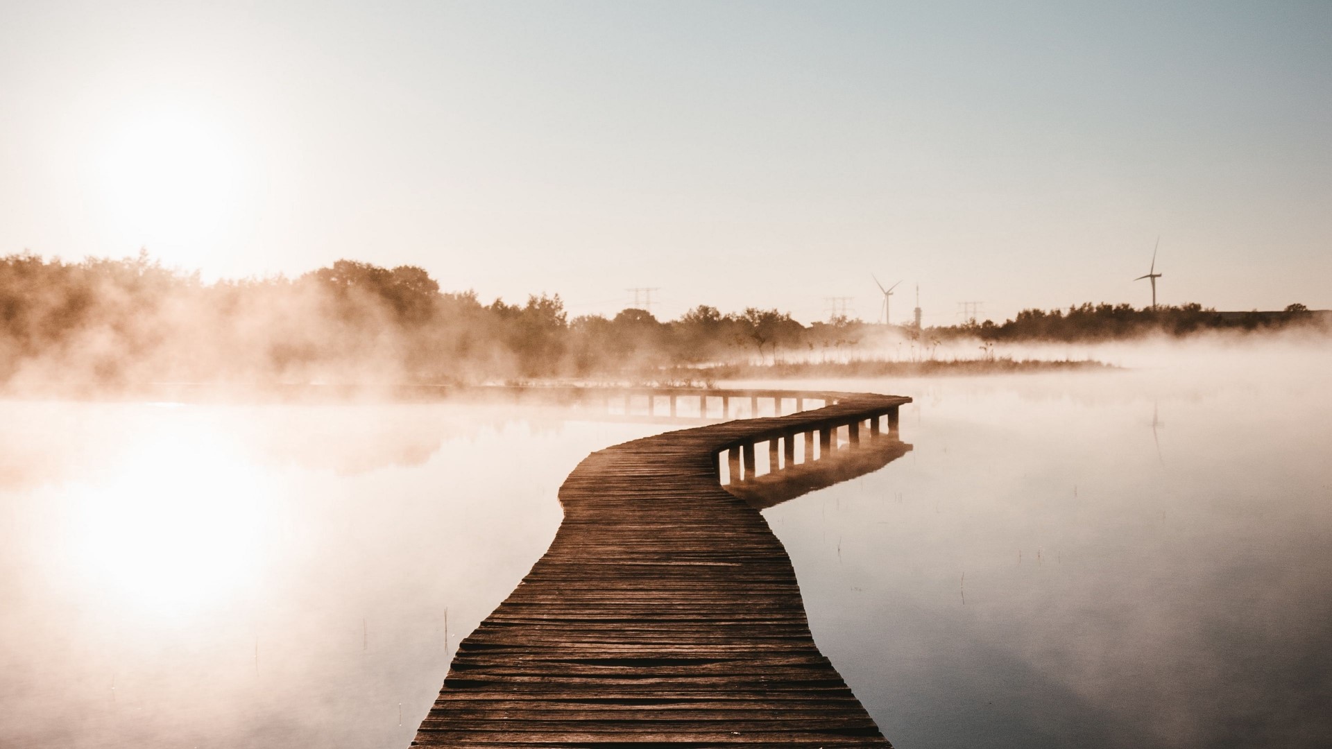 Wooden boardwalk over misty lake with wind turbines HD Wallpaper