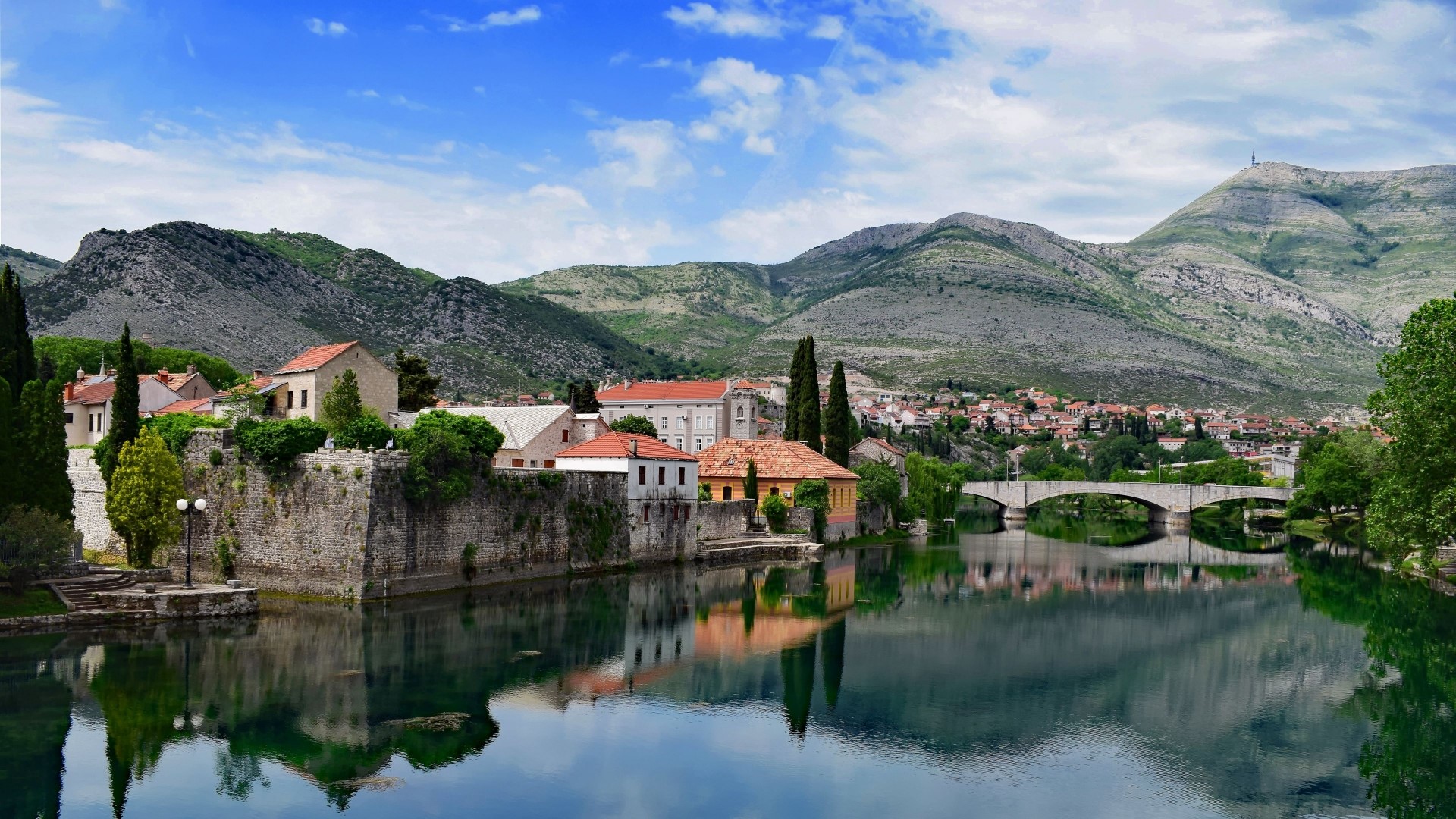 Stone buildings on a riverbank with mountains in the background HD Wallpaper