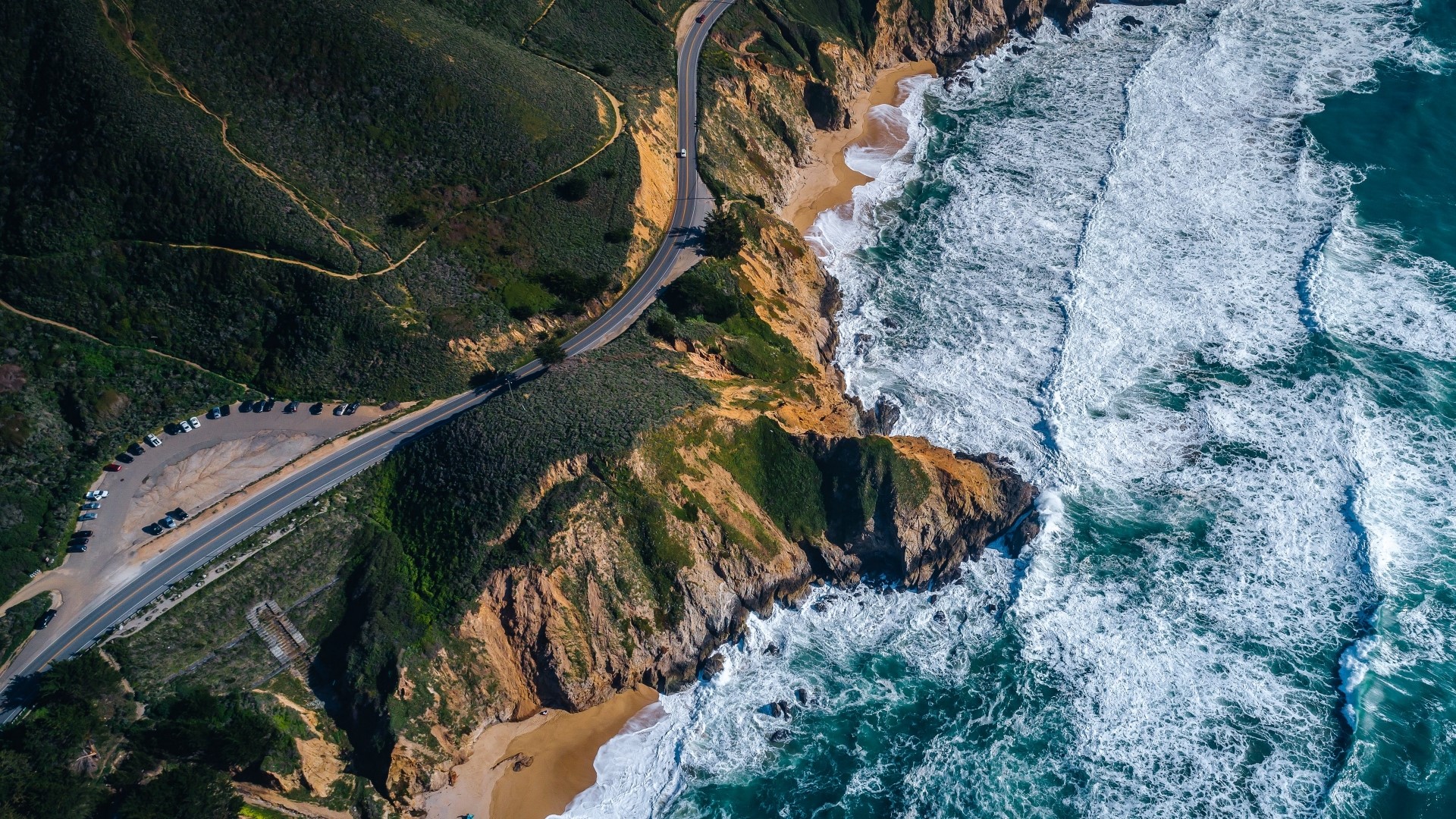 Aerial view of a winding road along a rugged coastline with crashing waves HD Wallpaper